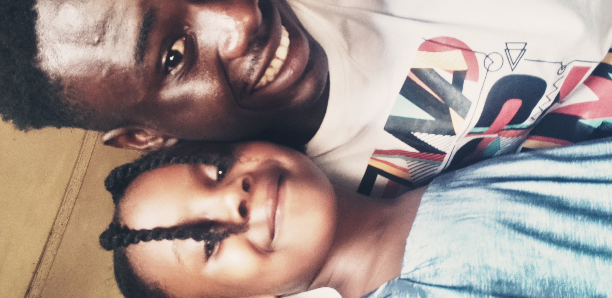 The image displays a close-up portrait of an adult male and a young child, captured in what appears to be an indoor setting in Narayi, Nigeria. The adult male, positioned on the right, is of African descent with short, dark hair and a faint mustache. He is smiling broadly, revealing his teeth, and wears a white t-shirt adorned with a multi-colored geometric pattern. The pattern includes various shapes and lines in red, black, yellow, teal, and white. The child, of African descent and appearing to be between three and seven years old, is situated to the left and slightly forward of the male. She has dark braided hair, with some braids falling across her forehead, and displays a closed-mouth smile. She is dressed in a light blue garment with vertical pleats or folds. Both individuals are oriented towards the viewer. The background is out of focus, suggesting a shallow depth of field, and presents as a light brown, possibly an interior wall, with a subtle horizontal architectural line visible in the upper portion.