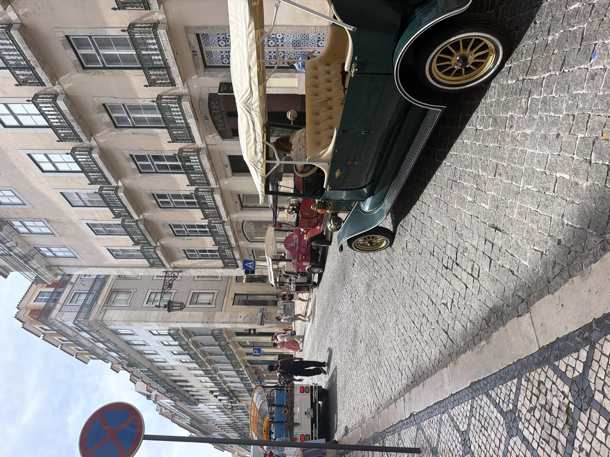 The image depicts a narrow, cobblestone street in Lisbon, Portugal, with tall, traditional European buildings lining both sides. In the foreground, a dark green vintage-style car with a light-colored canopy and tan upholstery is parked. The car has gold rims on its wheels. Further down the street, several other small open-air vehicles, some colored pink and yellow, are visible, suggesting they might be tourist vehicles. People are walking on the street and near the vehicles. The buildings feature multiple stories with balconies and ornate architectural details. A "no parking" sign is prominently displayed on the left side of the street. The sky is overcast with clouds. The overall scene suggests a bustling street in a historic urban area of Lisbon.