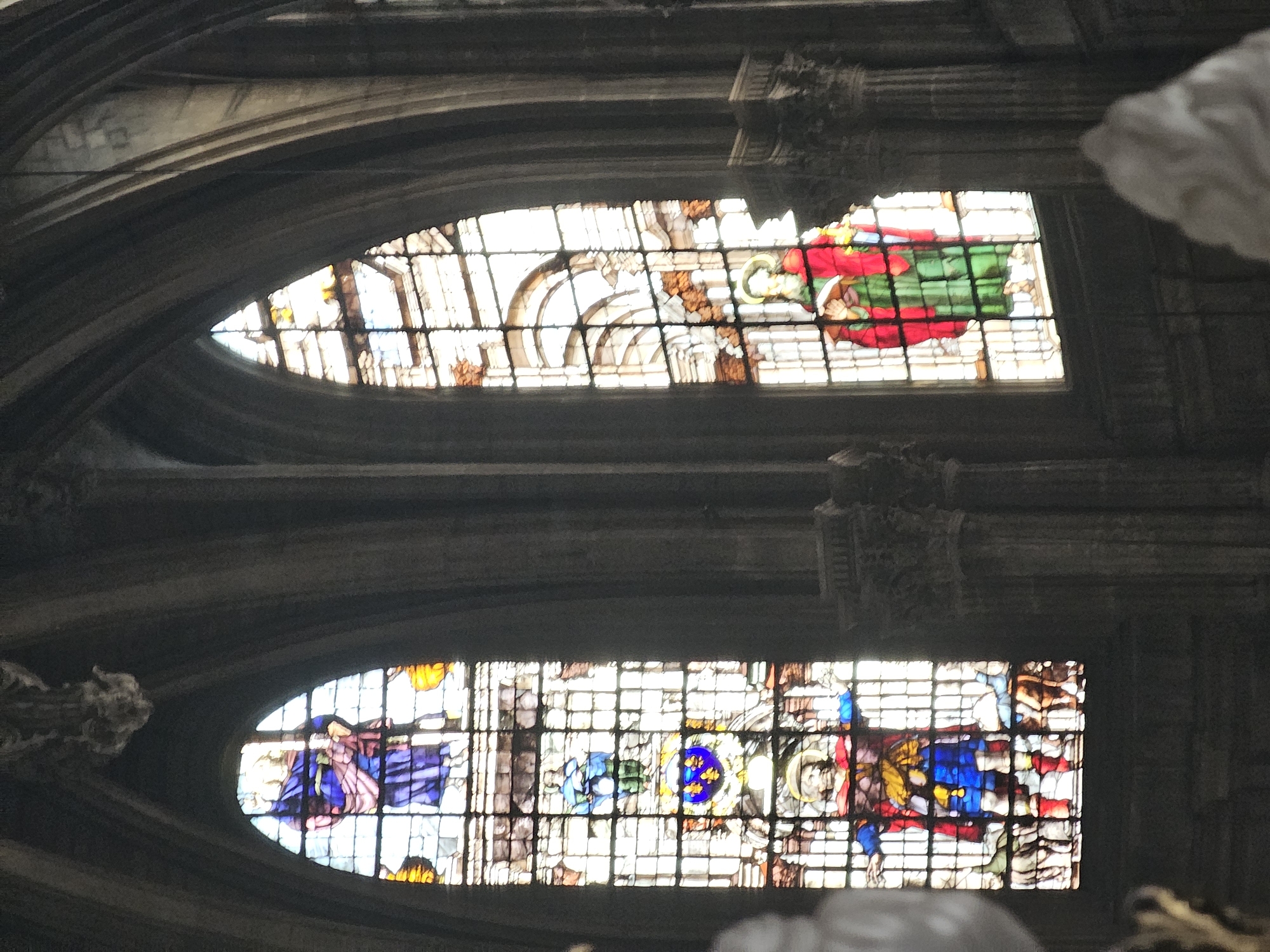 The image shows two stained-glass windows inside a Gothic-style building. The windows are arched and divided into small, grid-like panes. The lower window depicts a figure in blue and purple robes, a blue floral emblem, and another figure in red and blue. The upper window shows a figure in red and green robes. The architectural structure surrounding the windows appears to be made of dark stone with ornate carvings. The location is likely a church or cathedral, potentially in Paris, France, given the architectural style and stained glass artwork.