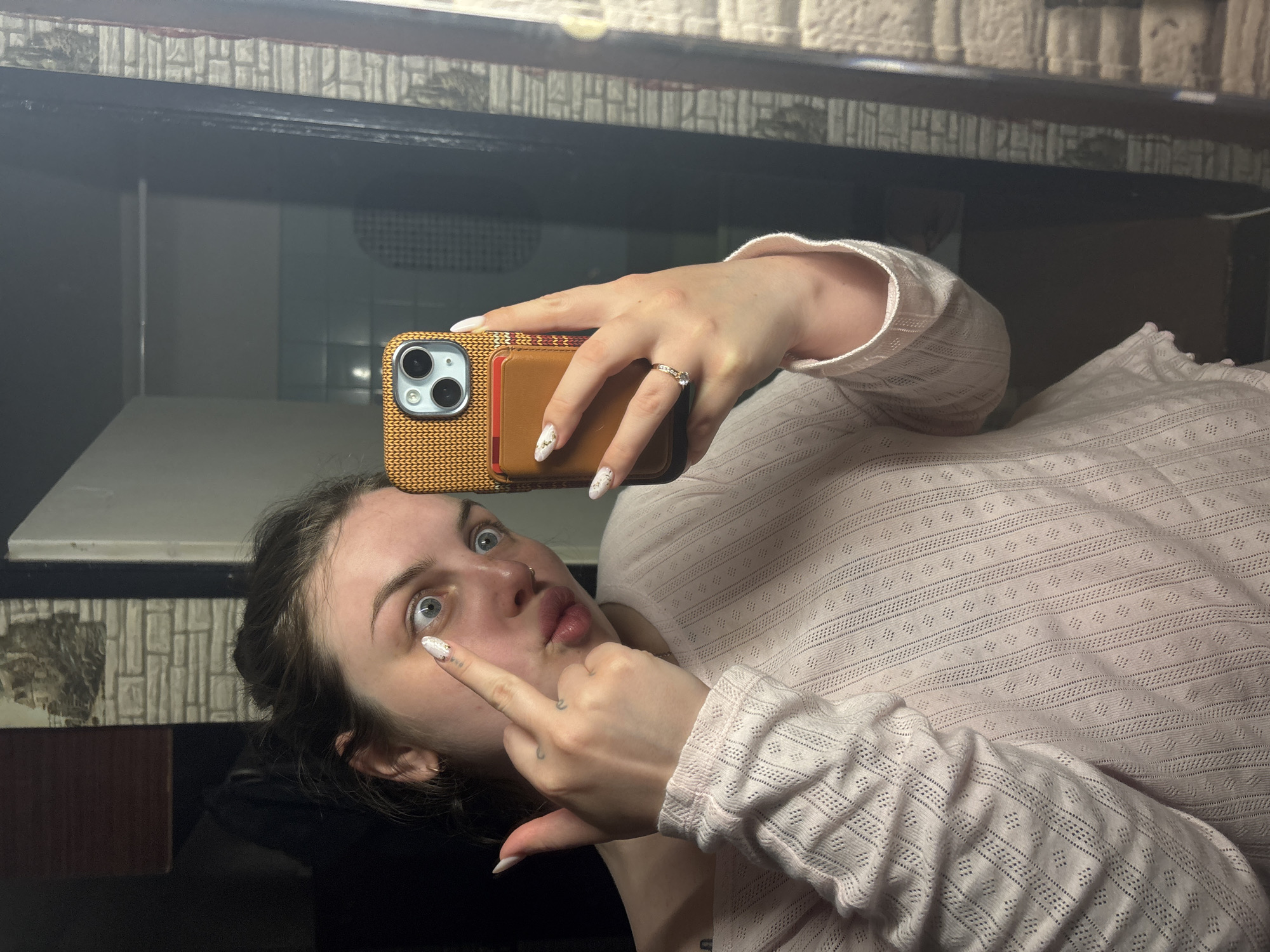A woman is taking a selfie in what appears to be a mirror. She is holding a phone with a brown case and is pointing at her eye with her left hand. She is wearing a light pink, long-sleeved, patterned shirt. Her hair is dark and pulled back. The background shows a room with tiled walls and a dark-colored shelf or ledge. The location cannot be determined, including the city of Chornomorskoe, Ukraine.