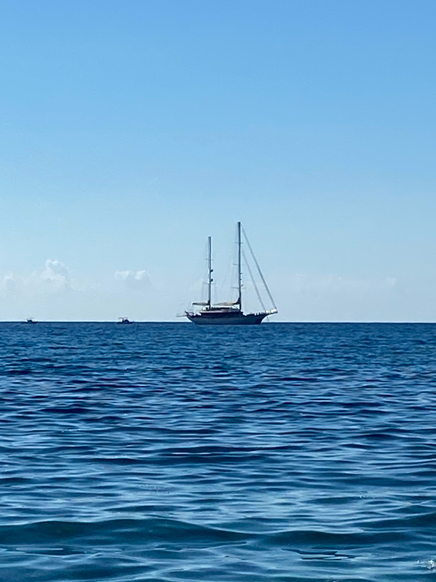 A wide shot captures a serene seascape under a clear, pale blue sky. The vast expanse of deep blue water fills the foreground and middle ground, with subtle ripples indicating gentle movement. On the horizon, a majestic sailing yacht with two tall masts and unfurled sails dominates the scene. To the left, two smaller boats are visible in the distance. The overall impression is one of tranquility and open water. The image is undated and has no explicit indications of its specific location in Prijevor, Montenegro, but it depicts a coastal or marine environment.