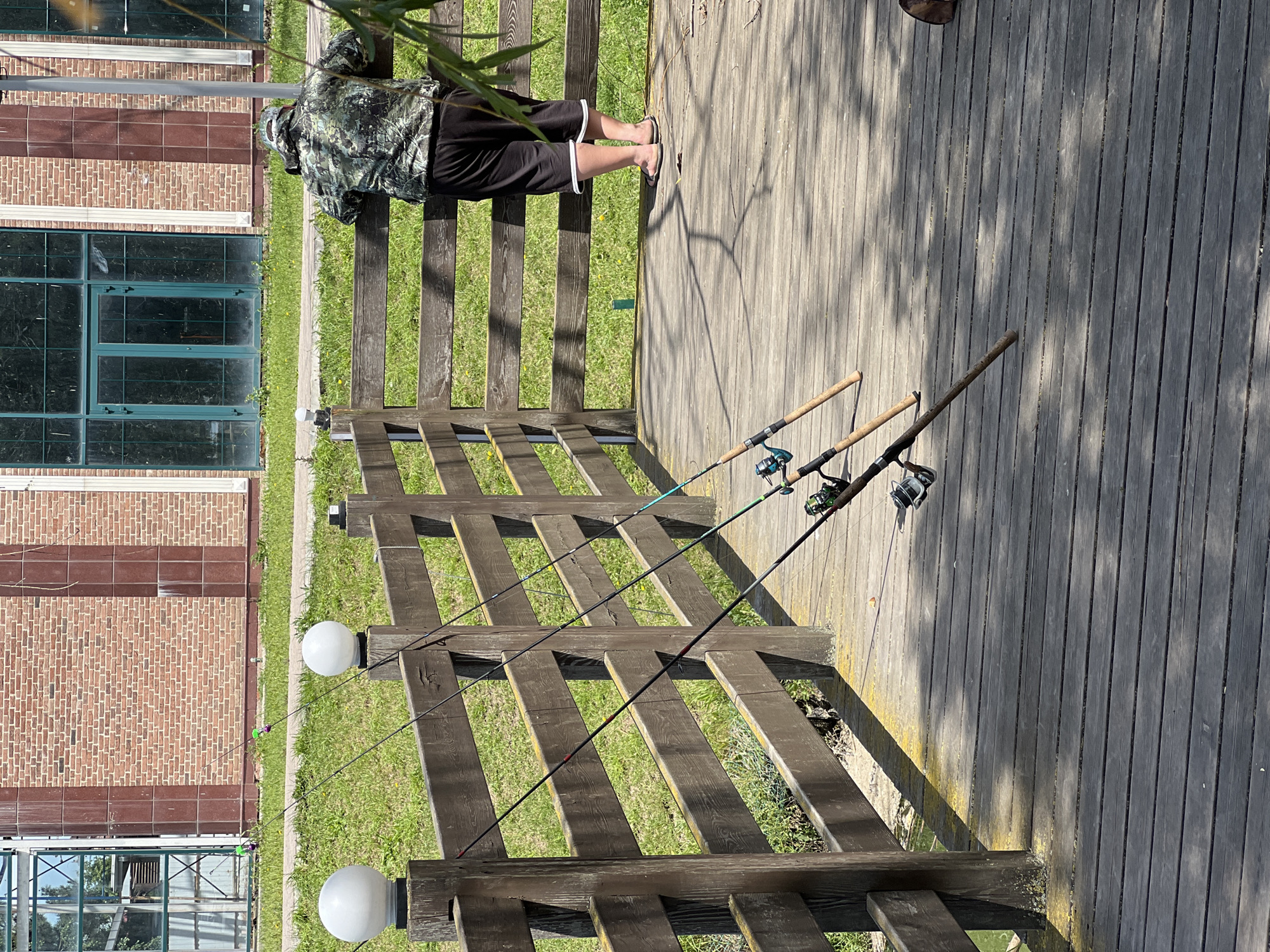 The image, taken in Krasnodar, Russia, captures an outdoor scene dominated by a wooden deck or pathway. A person, viewed from behind, stands on this wooden surface. They are wearing a camouflage-patterned long-sleeved shirt, black shorts with white trim, and flip-flops. Their posture suggests they are relaxed, possibly overseeing fishing rods.
Leaning against a sturdy wooden railing that borders the pathway are at least three fishing rods equipped with reels. These rods extend outwards, suggesting they are actively in use or set up for fishing. The wooden railing itself is robust, consisting of thick horizontal and vertical planks. Along the railing, two spherical white outdoor lights are visible on posts.
Beyond the railing and a strip of green grass, a large building with brick walls and numerous large, dark-framed windows is visible in the background. The brickwork features both reddish-brown and lighter-colored sections.
The scene is brightly lit by natural sunlight, indicated by the strong, distinct shadows cast across the wooden deck and the grassy area by the railing and some overhead foliage. The weather appears clear and sunny, likely during the day, either morning or afternoon. The overall atmosphere is calm, suggesting a leisurely outdoor activity like fishing. No visible text is present in the image.