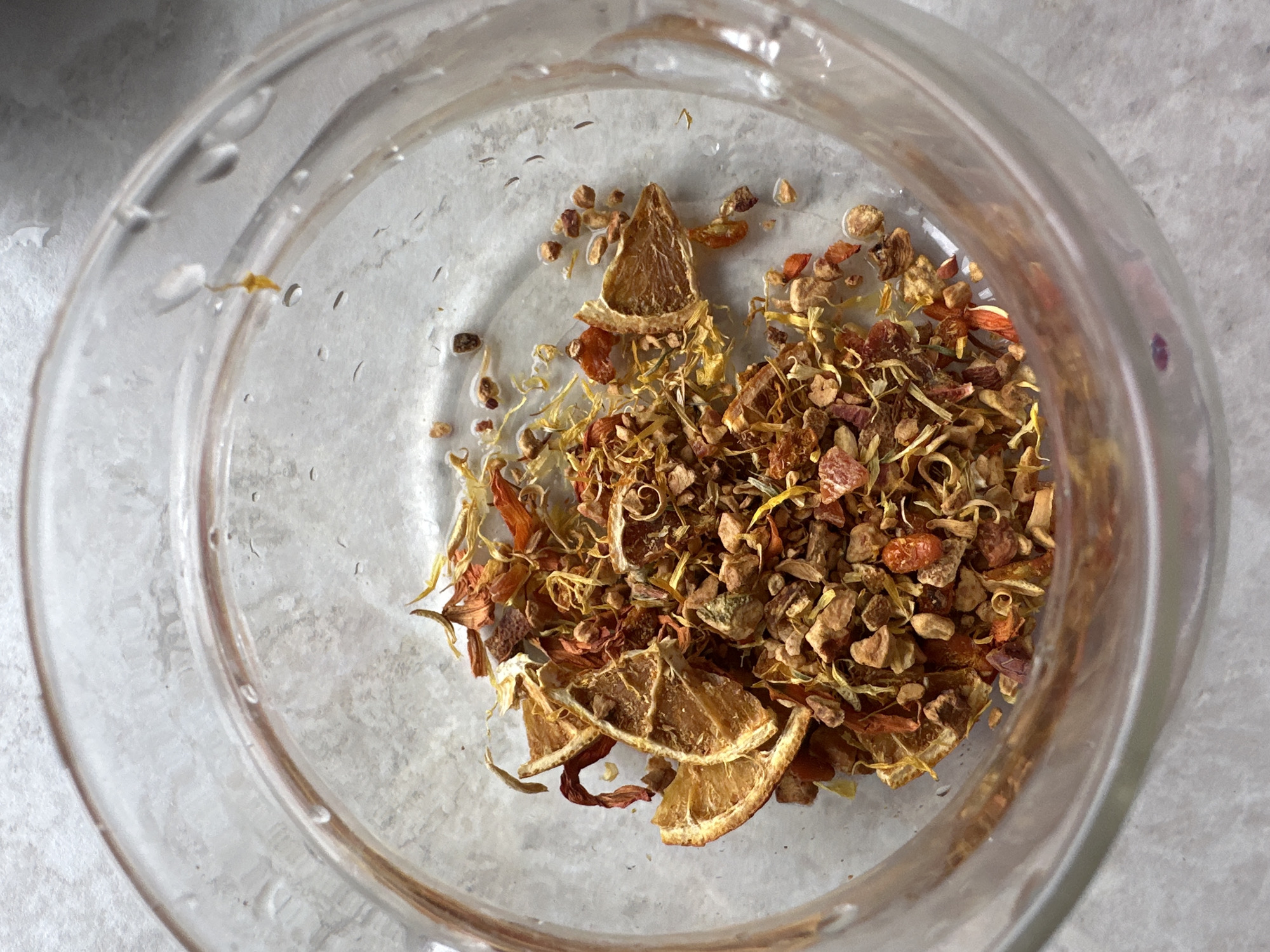 A top-down view shows a clear glass container filled with dried ingredients. The contents appear to be a mix of dried orange slices, various small brown and reddish-brown granules, and yellow thread-like pieces. The exterior of the glass container is slightly wet with visible droplets. The background is a light gray, possibly a counter or table surface.
There is no data regarding the image that provides the location in the city of Kharnavy, Russia.