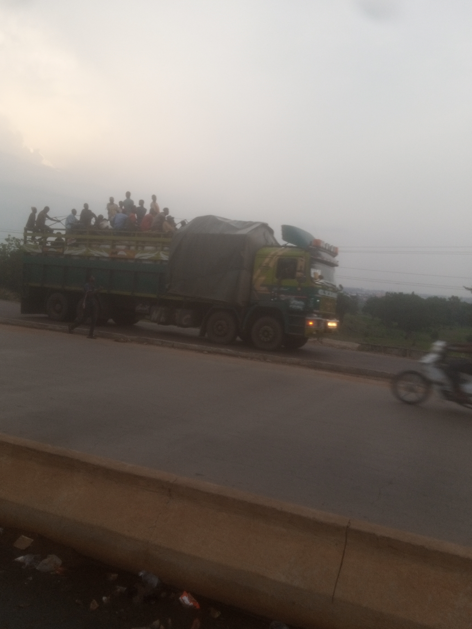 A low-angle, outdoor, medium shot shows a flatbed truck loaded with what appears to be produce, covered by a tarp, and carrying several passengers on top. The truck is green and lime green with yellow and orange lights on the front. It is on a paved road, with a concrete barrier along the right side. A person in dark clothing is walking in front of the truck. A motorcycle is moving away from the truck on the road. The sky is overcast and the background shows trees and a distant city. The scene is lit by daylight. The location is identified as Goni Gara, Nigeria.
