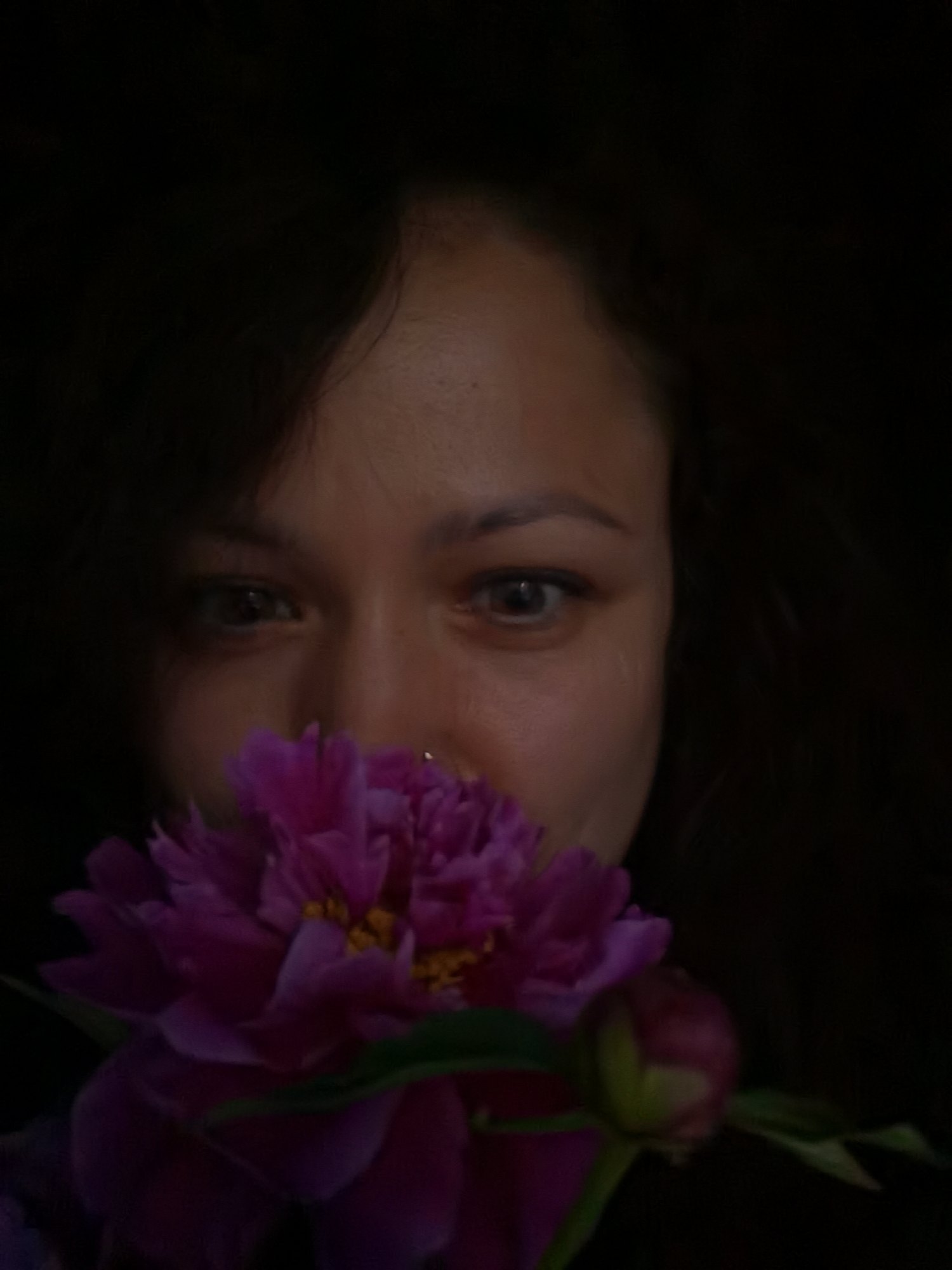 The photograph, taken in Chaikovsky, Russia, features a close-up shot of a person's face partially obscured by a vibrant pink peony. The person's eyes are visible and appear to be looking directly at the camera. They have dark hair and what seems to be a nose piercing. The lighting is low, creating a dramatic effect with the dark background emphasizing the flower and the face. The peony is in full bloom, with delicate petals and yellow stamens at its center. A green stem and leaves are also visible, along with a small, unopened bud to the right of the main flower. The image has a soft focus, adding to its artistic quality.