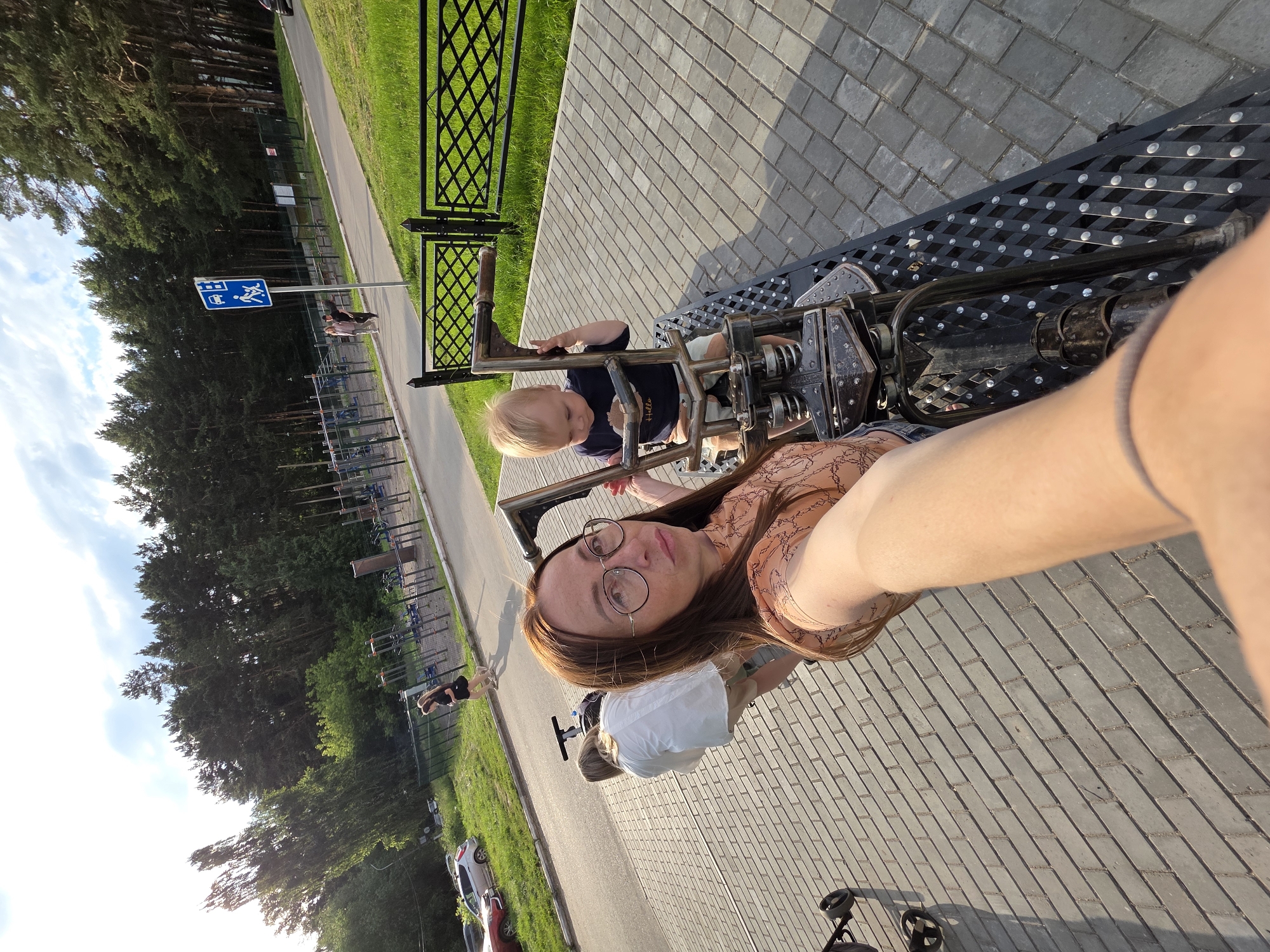 This is a selfie taken outdoors in Chaikovsky, Russia. The image shows a woman with glasses and brown hair, wearing a patterned blouse, and a child with blonde hair wearing a dark blue shirt. They are on a metal structure, possibly part of an outdoor gym or playground equipment, with a cobblestone path and a grassy area visible. A stroller is also present. In the background, there are trees, a fence, and a road with cars. A blue sign with pictograms is also visible near the road. The lighting suggests it is daytime, likely late afternoon or early evening, with the sun casting long shadows. The overall mood appears casual and relaxed, suggesting a family outing. The text on the child's shirt is "Hello."
