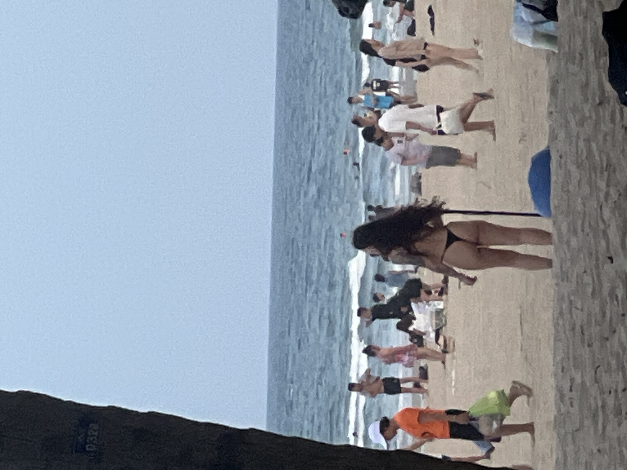 The image captures a bustling beach scene in Đà Nẵng, Vietnam. A large group of people are enjoying the sand and surf. Several individuals are visible in the water, while others are scattered across the beach. One woman in the foreground is wearing a black bikini and appears to be posing or stretching. Many people are dressed in casual beach attire, including swim trunks and t-shirts.
The activity appears to be a casual gathering or outing at the beach, with some people engaged in what looks like a group exercise or activity. The weather seems to be overcast, with a pale blue sky and calm ocean waves. The sand is light brown and appears somewhat wet near the water's edge. The overall atmosphere suggests a relaxed and active day at the beach.
In the lower left corner of the image, a dark object, possibly a tree trunk or debris, is visible. Faint text, "0328", is discernable on this object. The image is oriented vertically, with the ocean on the left and the beach on the right. The composition suggests the photo was taken from a slightly elevated position, looking down at the beachgoers.