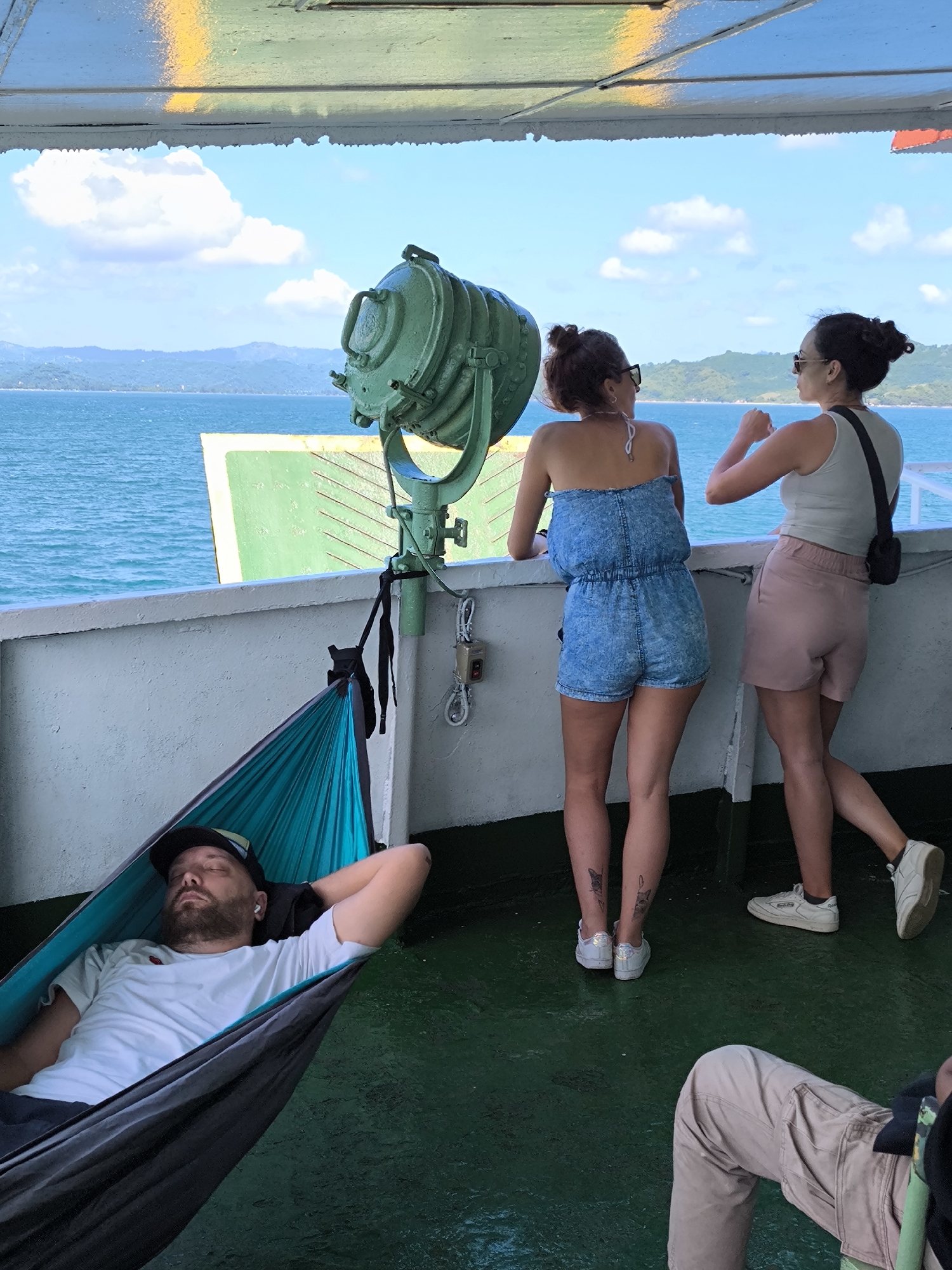 The image captures a vibrant daytime scene aboard a ferry or large boat, likely in a tropical coastal region. The setting features clear blue water with distant green, hilly islands under a bright blue sky with scattered white clouds.
In the foreground, a man is comfortably resting in a teal and black hammock, eyes closed, suggesting he is asleep or relaxing. He has a beard, is wearing a white t-shirt, a black baseball cap, and white earbuds. The hammock is secured to the boat's white wall.
Two women are standing at the boat's railing on the right side of the frame, looking out towards the sea and the landscape. The woman on the left is wearing a strapless, light blue denim-look romper and sunglasses perched on her head; she has dark hair styled in a bun and visible tattoos on her calves. The woman on the right is wearing a light-colored tank top, pink/beige shorts, sunglasses, and has dark hair also in a bun, with a black cross-body bag. Both appear engaged in observing their surroundings. Partially visible in the bottom right corner are the legs of a fourth person, wearing light brown pants.
A prominent feature in the center is a large, green industrial-style spotlight or searchlight, mounted on a pole and angled towards the left. The boat's deck is a distinctive green color and appears somewhat wet or reflective. The ceiling above, visible at the top of the image, is painted green and yellow. Part of another boat structure, possibly a ramp or another section of the same ferry, is seen in the water to the left, also green and yellow. An electrical control box with red and white buttons is mounted on the wall near the hammock.
The overall atmosphere suggests a leisurely journey under sunny weather. No visible text is clearly legible in the image. The scene aligns with a coastal setting in Indonesia.