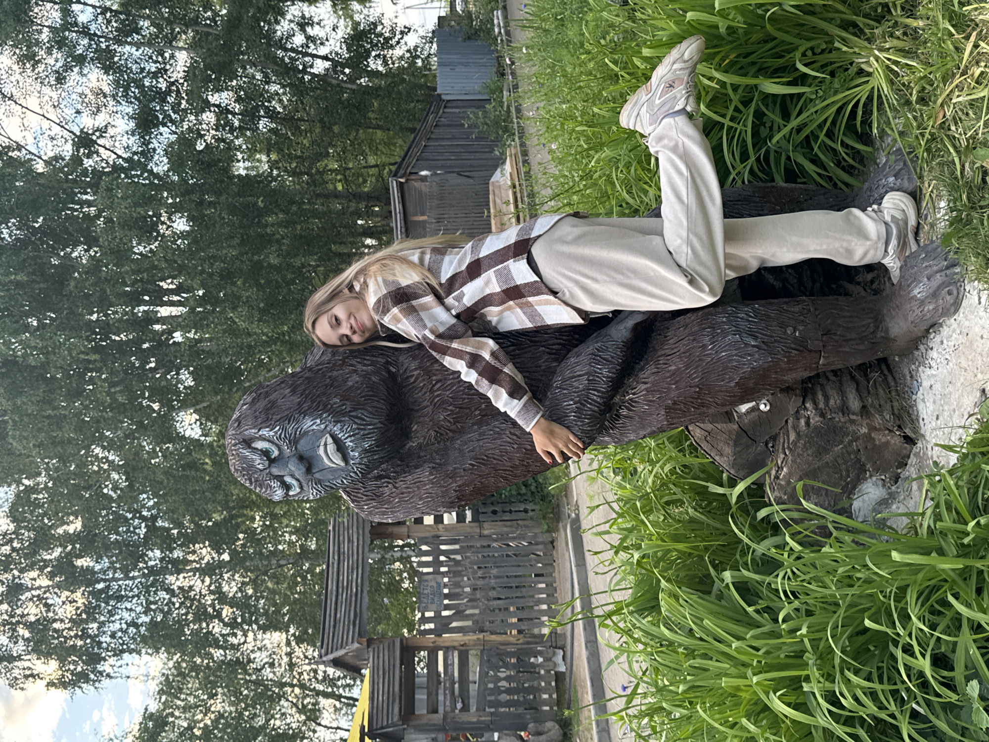A young woman with blonde hair and fair skin is posing for a photo outdoors. She is wearing a plaid shirt with brown, white, and beige squares, and light-colored sweatpants. She is leaning against a large, dark brown statue of a gorilla. Her left arm is wrapped around the gorilla's torso, and her right leg is bent and extended backward. She is smiling at the camera.
The background of the image shows a park-like setting with lush green grass and tall trees. There are wooden fences and structures visible behind the gorilla statue, suggesting an outdoor recreational area or a park exhibit. The sky is partly cloudy. The specific location appears to be a park or an attraction in the city of Chaikovsky, Russia, given the presence of wooden folk-style structures and the general environment.