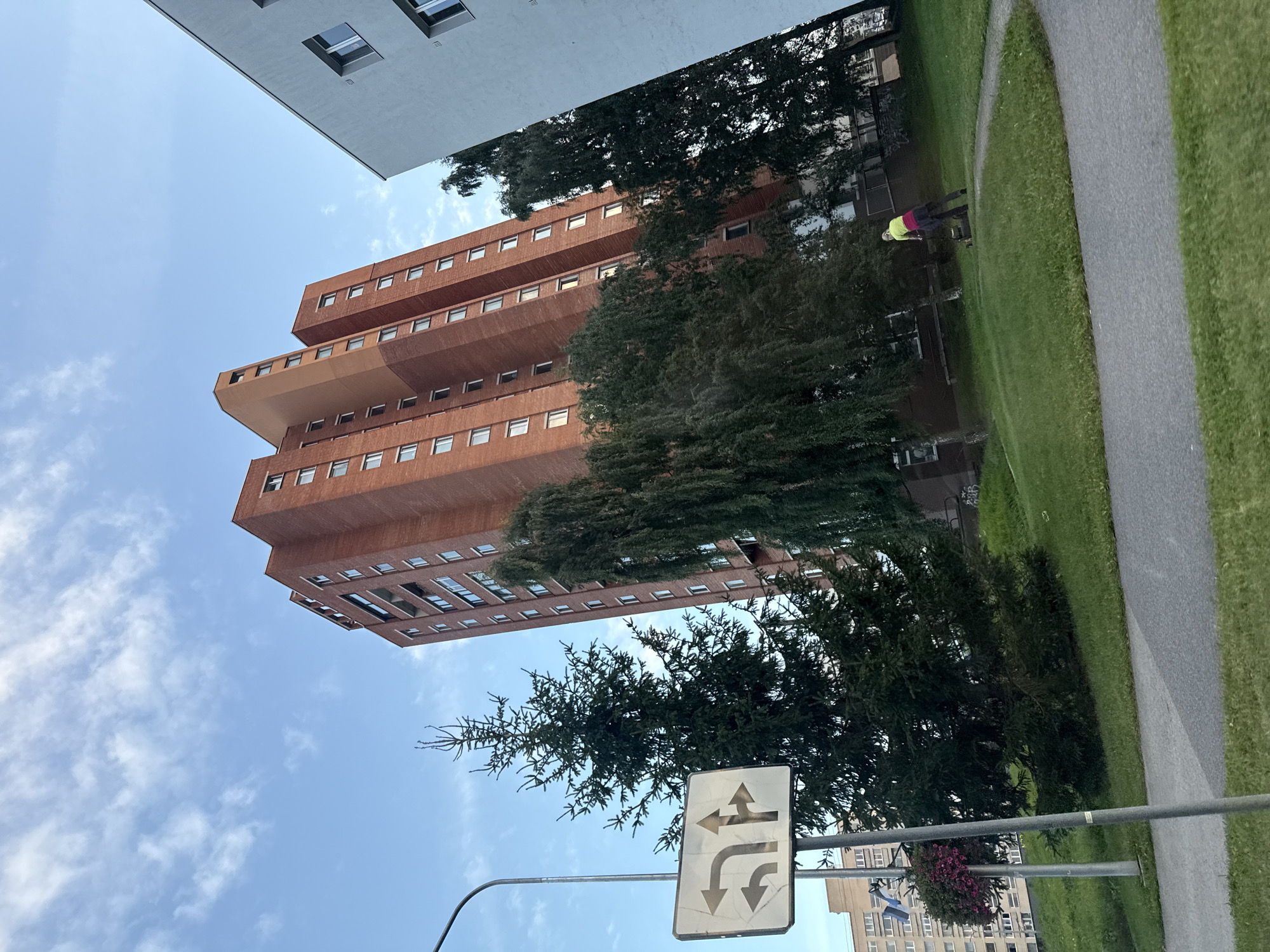 The image captures a daytime scene in Tallinn, Estonia, with a focus on a tall, red brick apartment building. To the left, a traffic sign indicating turning lanes is visible next to a tall evergreen tree. A person is seen in the mid-ground operating a lawnmower on a grassy area. Another modern, light-colored building is partially visible on the right side of the frame. The sky is blue with scattered clouds.