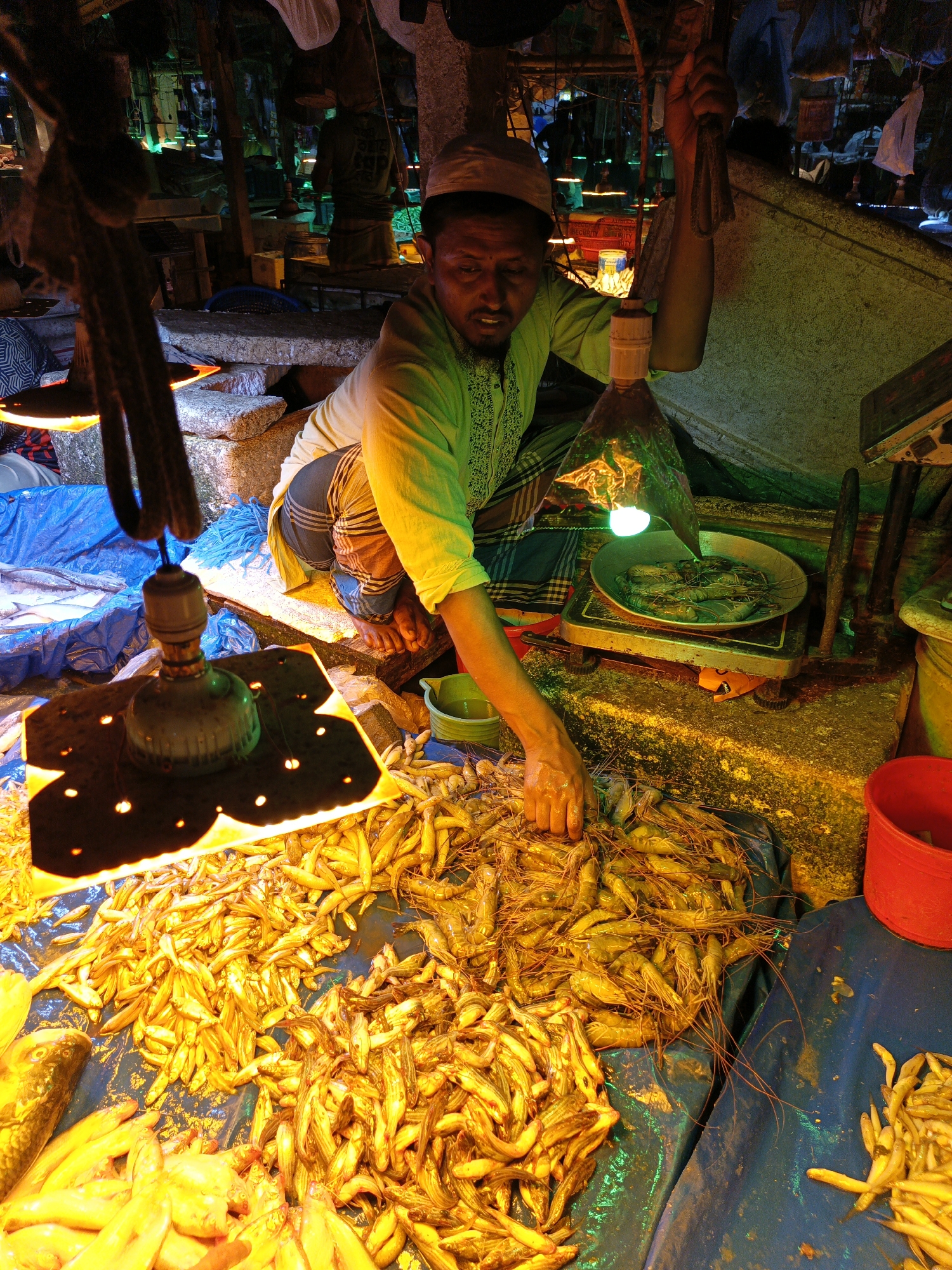 A fishmonger in Dhaka, Bangladesh, kneels at his stall sorting small, pale-yellow fish and shrimp under bright electric lights. He wears a light green embroidered shirt and a white skullcap. He is barefoot. To his right, a pan of larger shrimp sits on a scale. The stall has blue tarps and is surrounded by concrete blocks. The scene is in a dimly lit market.
