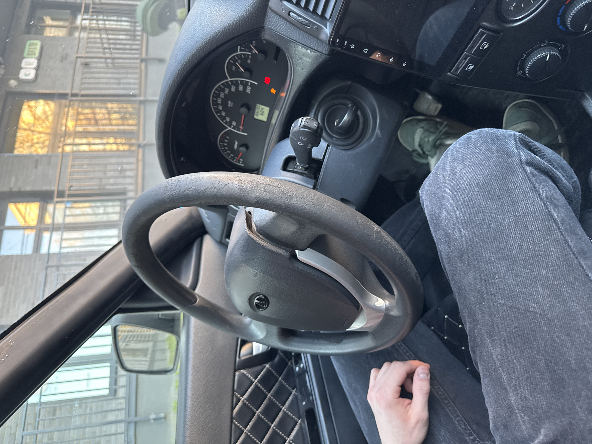 A view from inside a car, likely in Perm, Russia, based on the dashboard and interior design common in Russian vehicles. The driver, wearing green shoes and grey jeans, is visible. A steering wheel with a Russian car emblem (possibly UAZ) dominates the foreground. The dashboard includes a speedometer showing approximately 60 km/h. The passenger-side interior has a diamond-stitched pattern. Through the windshield, a multi-story building and fire escape are visible. The ignition is on.