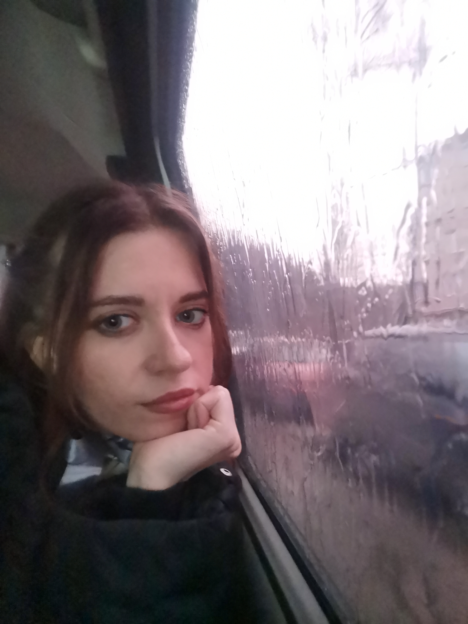 A woman is sitting on a vehicle next to a window covered with rain, looking out. The location is Unknown, Unknown. The woman has brown hair and is wearing a black top. She is resting her chin on her hand. Outside the window, the blurred cityscape is visible through the rain streaks.
