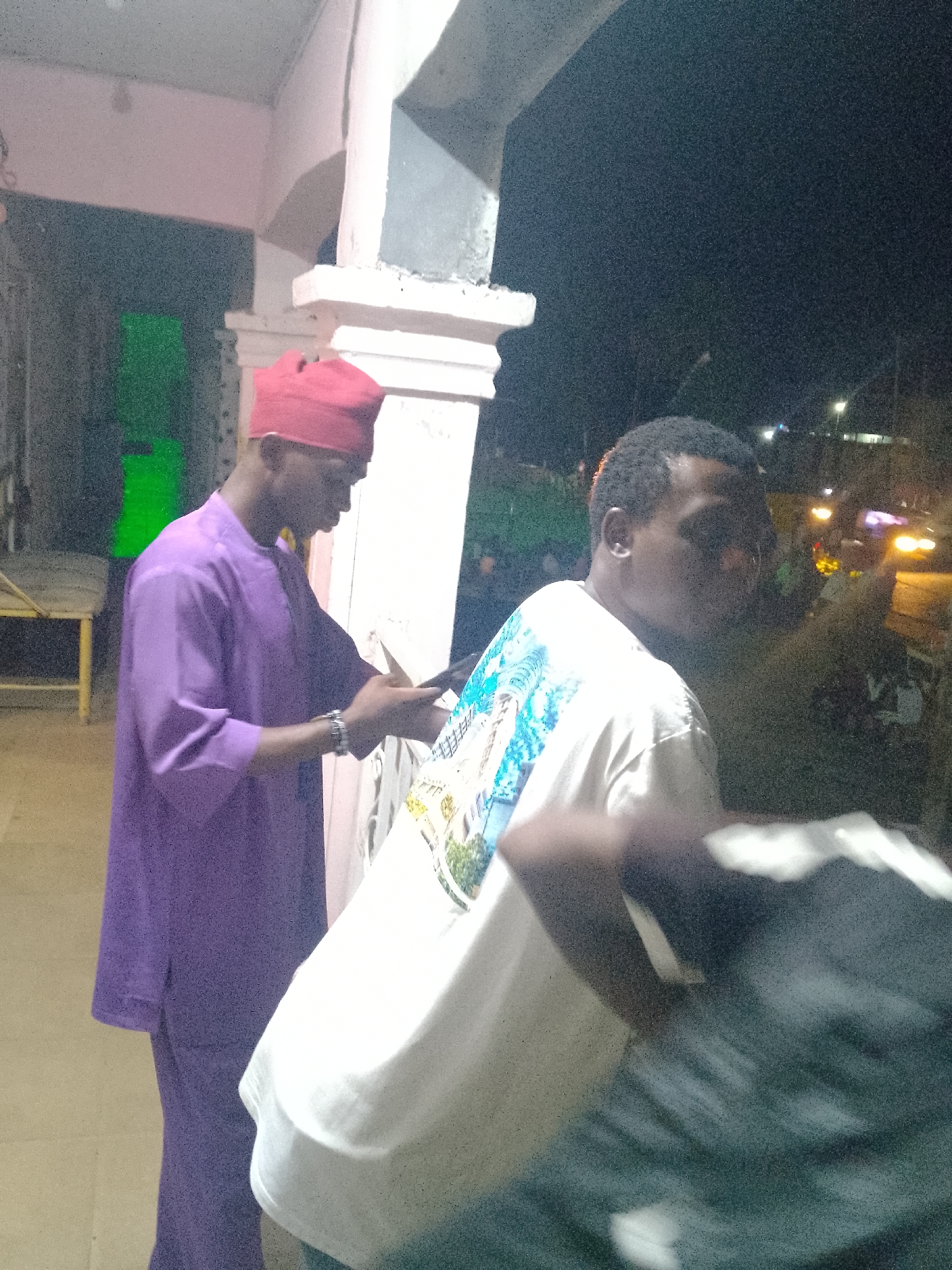 The image depicts an urban night scene in Jalingo, Nigeria, featuring two male subjects and elements of a building and street environment. In the foreground, a dark-skinned male is shown from the back, turned slightly to his right, revealing a white short-sleeved t-shirt with a detailed graphic print on the back depicting a landscape with buildings and foliage. His head is turned to his right, partially obscuring his face.
In the mid-ground, a second dark-skinned male is standing in profile, facing right. He wears a red cap and a purple traditional tunic and trousers. He is engaged with a smartphone, holding it with both hands and looking down at the screen. A metallic bracelet is visible on his left wrist.
The immediate background behind the man in purple consists of a light-colored building facade with structural elements resembling pillars or columns. A dark opening or recessed area to the left of the purple-clad man contains a prominent, illuminated green rectangular object. Further to the left, a portion of a yellow structure, possibly a bench or table, is visible. The distant background beyond the foreground subject reveals a dark night sky dotted with multiple artificial light sources, including bright streetlights and blurred yellow and red lights from vehicles, indicating a public street or thoroughfare. The overall scene is dimly lit, resulting in a grainy image quality.