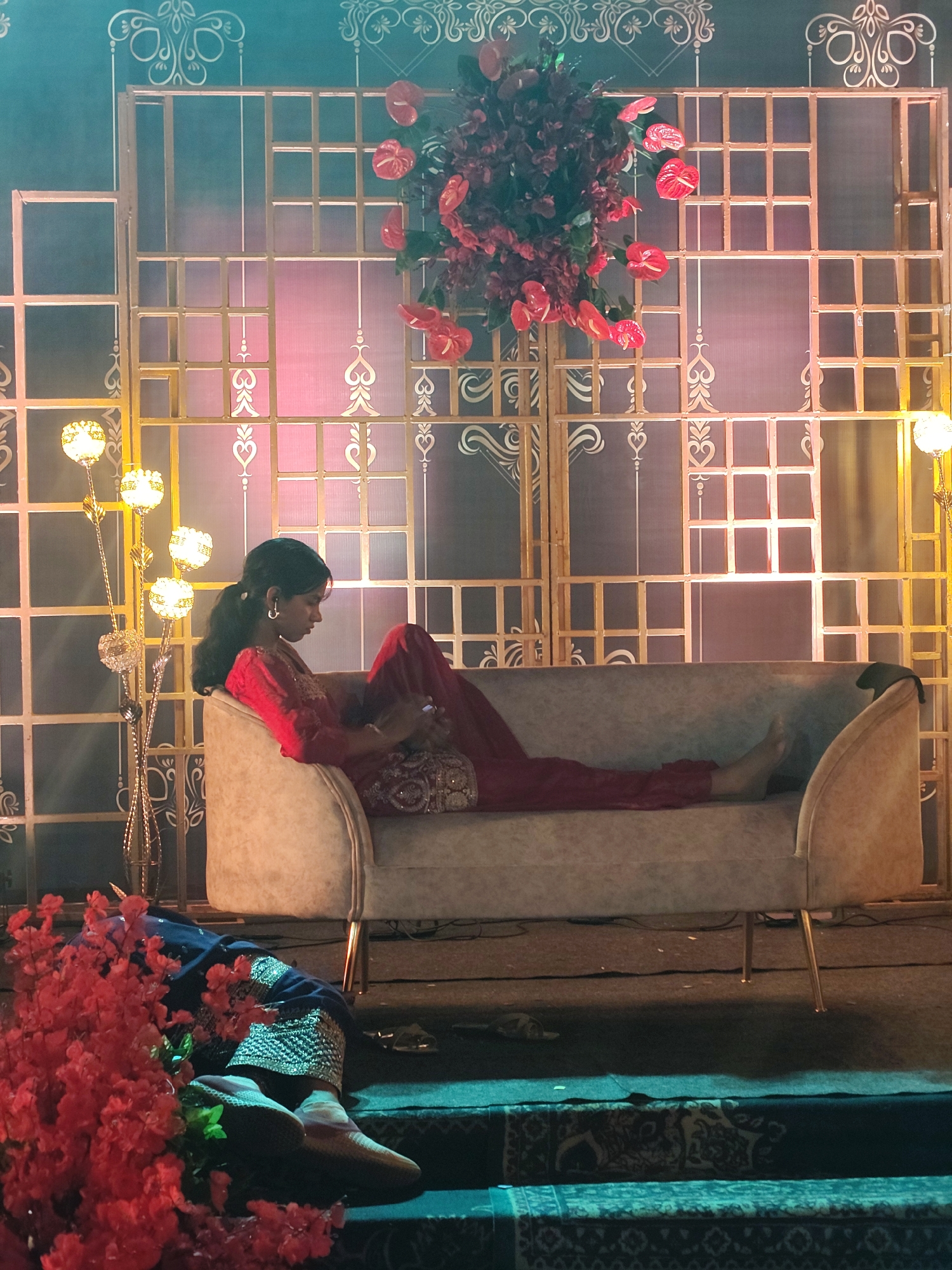 The image depicts a woman reclining on an upholstered sofa at an evening event in Kalamassery, India. She is wearing a red traditional outfit, possibly a salwar kameez, with decorative sleeves and trousers, and her dark hair is pulled back in a ponytail. She is looking down at a device held in her hands, appearing to be engaged with a mobile phone. Her posture is relaxed, with one bare foot visible.
The background features an elaborate decorative backdrop. It consists of golden grid-like panels overlaid on a surface that shifts in color from a dark teal-green at the top left to pinkish-purple in the center, illuminated by artificial lights. White and golden intricate patterns, including heart shapes and swirls, are visible on the backdrop. A large arrangement of red flowers, including anthuriums, hangs from the top center of the backdrop.
To the left of the woman stands a tall, ornate floor lamp with multiple spherical, glowing lights. Another similar lamp is partially visible on the far right. The sofa is a light-colored, possibly beige or cream, upholstered piece with slender golden legs. The floor beneath is dark, covered with patterned rugs, suggesting a raised platform or designated seating area. In the bottom left foreground, part of another person's legs, dressed in dark blue attire with silver embroidery and footwear, is visible, partially obscured by a cluster of red decorative flowers. A slipper is also seen on the ground near the sofa. The scene conveys a festive and formal atmosphere, typical of a social gathering or celebration. No text is visible in the image.
