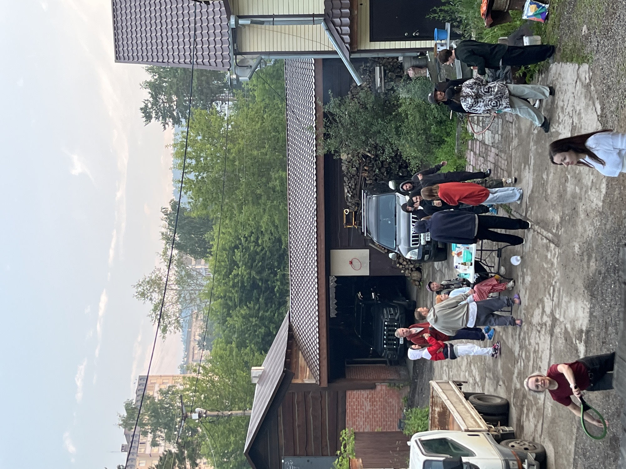 In Divnogorsk, Russia, on a cloudy day, a group of people are gathered in a yard with a residential building and a wooden structure with a garage. In the foreground, a woman in a red shirt and dark pants is holding a tennis racket. Several other individuals are interacting and standing around a table with drinks and food. Two SUVs are parked in the driveway, and a white truck is visible on the left. A pile of firewood is stacked near the garage. The overall scene suggests a casual outdoor gathering.