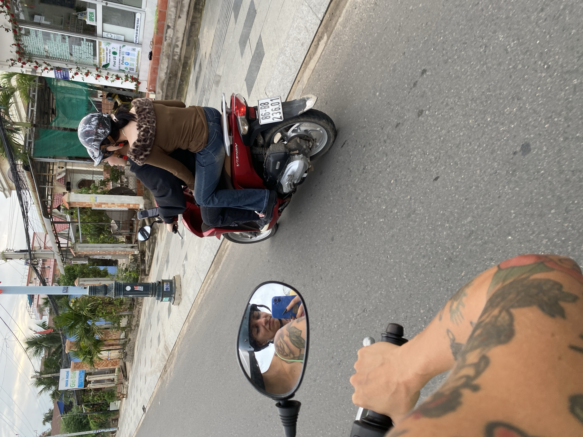 The image shows a first-person perspective of a person on a scooter. The rider has heavily tattooed arms. In the scooter's side mirror, the rider can be seen taking a selfie with a blue smartphone. Ahead, another person, wearing a helmet and tan jacket with a faux fur collar, rides on a red Honda Vision scooter. The license plate of the red scooter reads "86-B8 236.01". The scene takes place on a city street in Phu Thuy Ward, Vietnam. Street signs and businesses are visible on the side of the road, including a sign for a guesthouse called "Gió Bie" and a business with an "OPEN" sign.