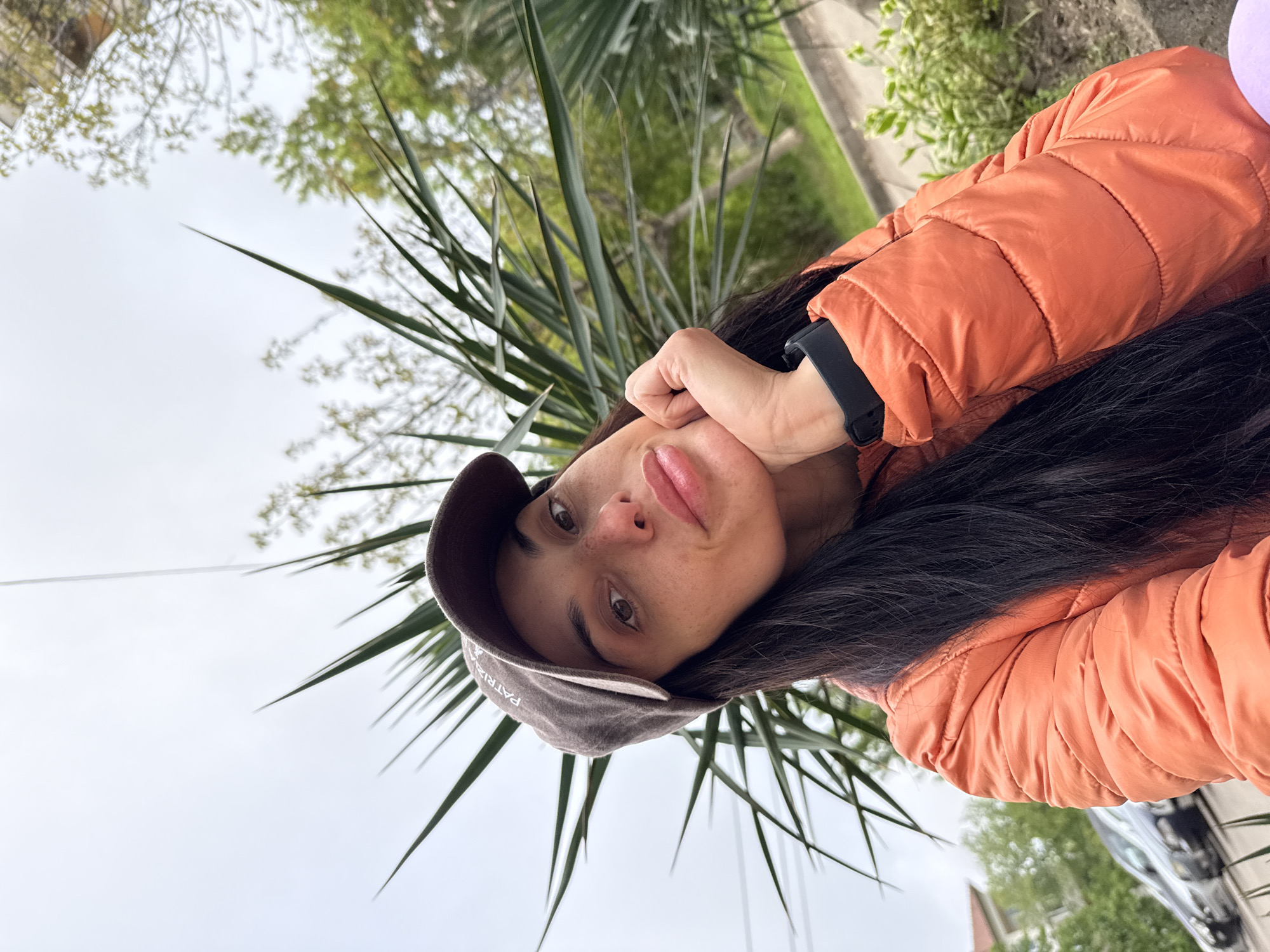 A woman with dark hair and a brown baseball cap is wearing an orange quilted jacket. She is holding her hand to her face. She appears to be standing outside near some greenery. The location is not identifiable as Pleven, Bulgaria.