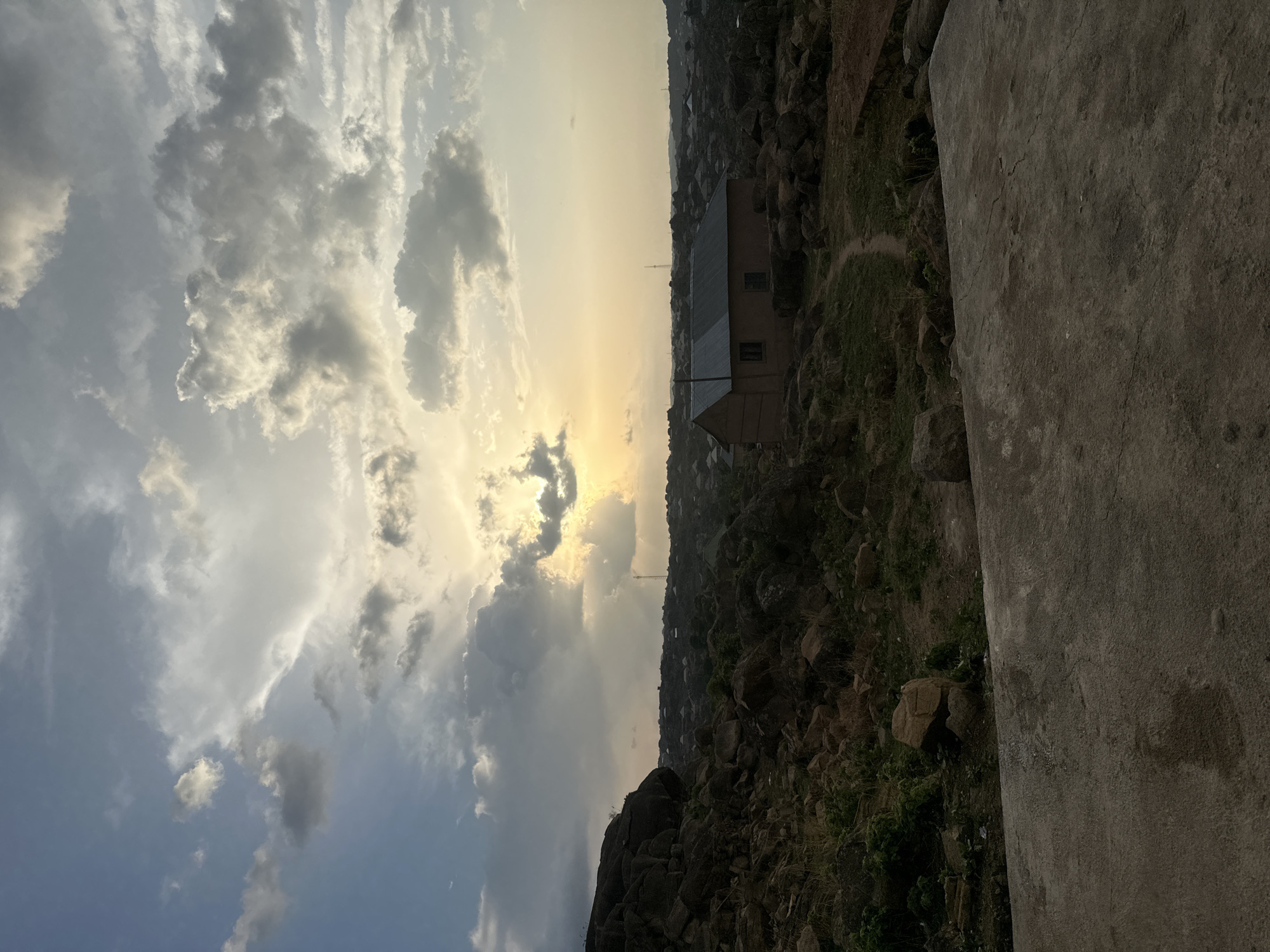 The image shows a landscape in Jos, Nigeria, featuring a sky filled with cumulus clouds, illuminated by a setting sun. The sunlight is visible between the clouds. Below the sky, the terrain is rocky and uneven, with scattered vegetation. A building is situated amid the rocks and vegetation. The foreground includes a rough, gray stone or concrete surface.