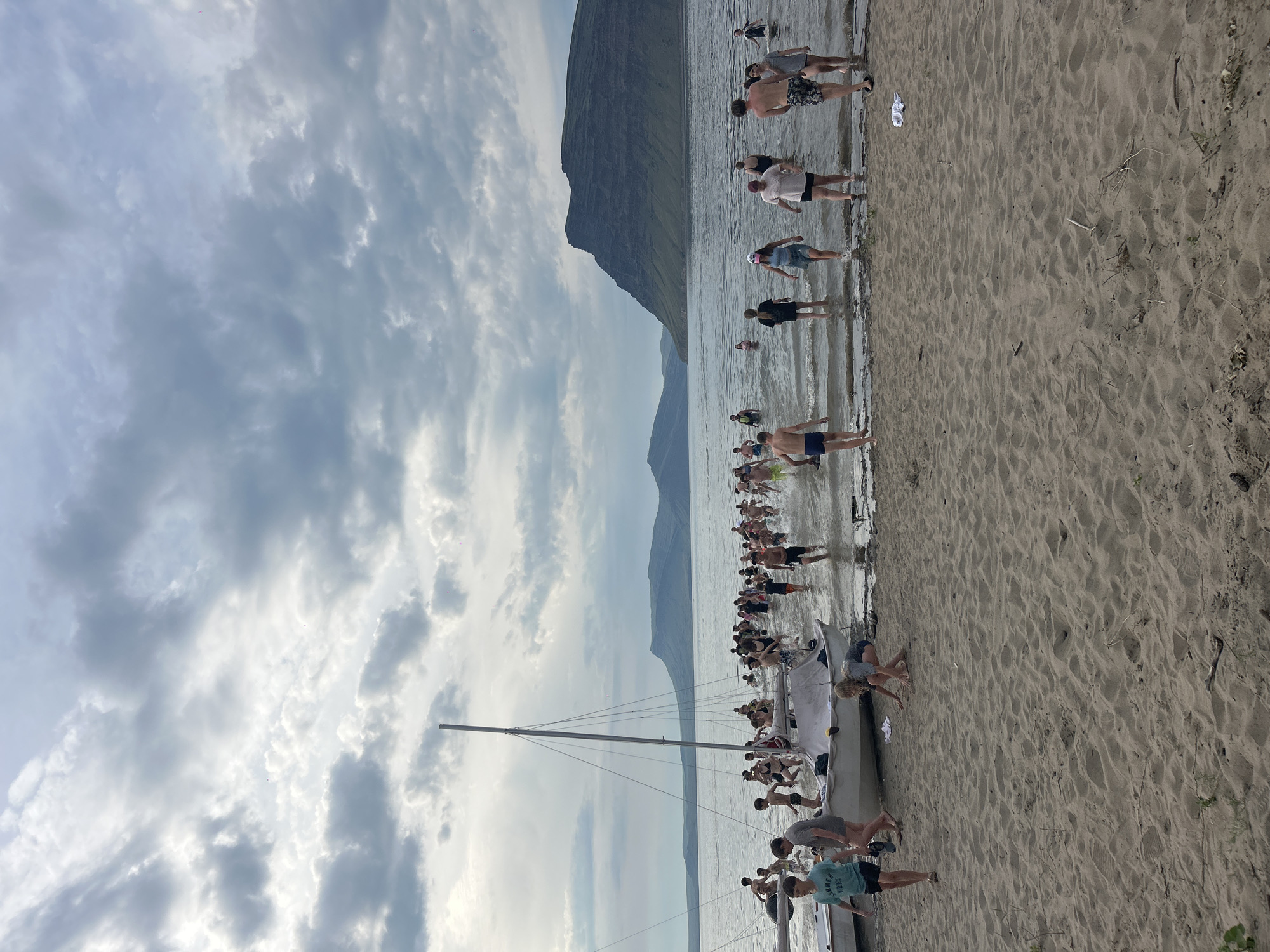 The image depicts a group of people, many of whom appear to be children and young adults, entering the water at a beach. There are two small sailboats partially visible on the sand and in the shallow water. The environment suggests a temperate climate with a cloudy sky and calm water. In the background, large, dark, flat-topped mountains rise from the horizon, framing the scene. The overall impression is of a recreational gathering or swimming activity taking place near the water's edge.