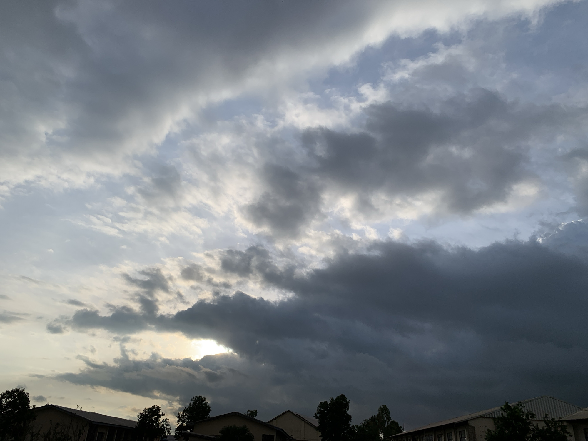 The image shows a dramatic sky over a section of the city of Kuru Karama, Nigeria. Heavy, dark clouds dominate the upper portion of the frame, with breaks of lighter clouds and hints of blue sky peeking through. The sun is partially obscured by the thick cloud cover, creating a diffused, somewhat ominous lighting condition. In the lower part of the image, the rooftops of several buildings are visible, silhouetted against the sky. These appear to be low-rise structures, possibly residential or administrative buildings, with trees interspersed between them. The overall impression is of a sky before or during a storm, with a somber and weighty atmosphere. There are no people or specific actions depicted.