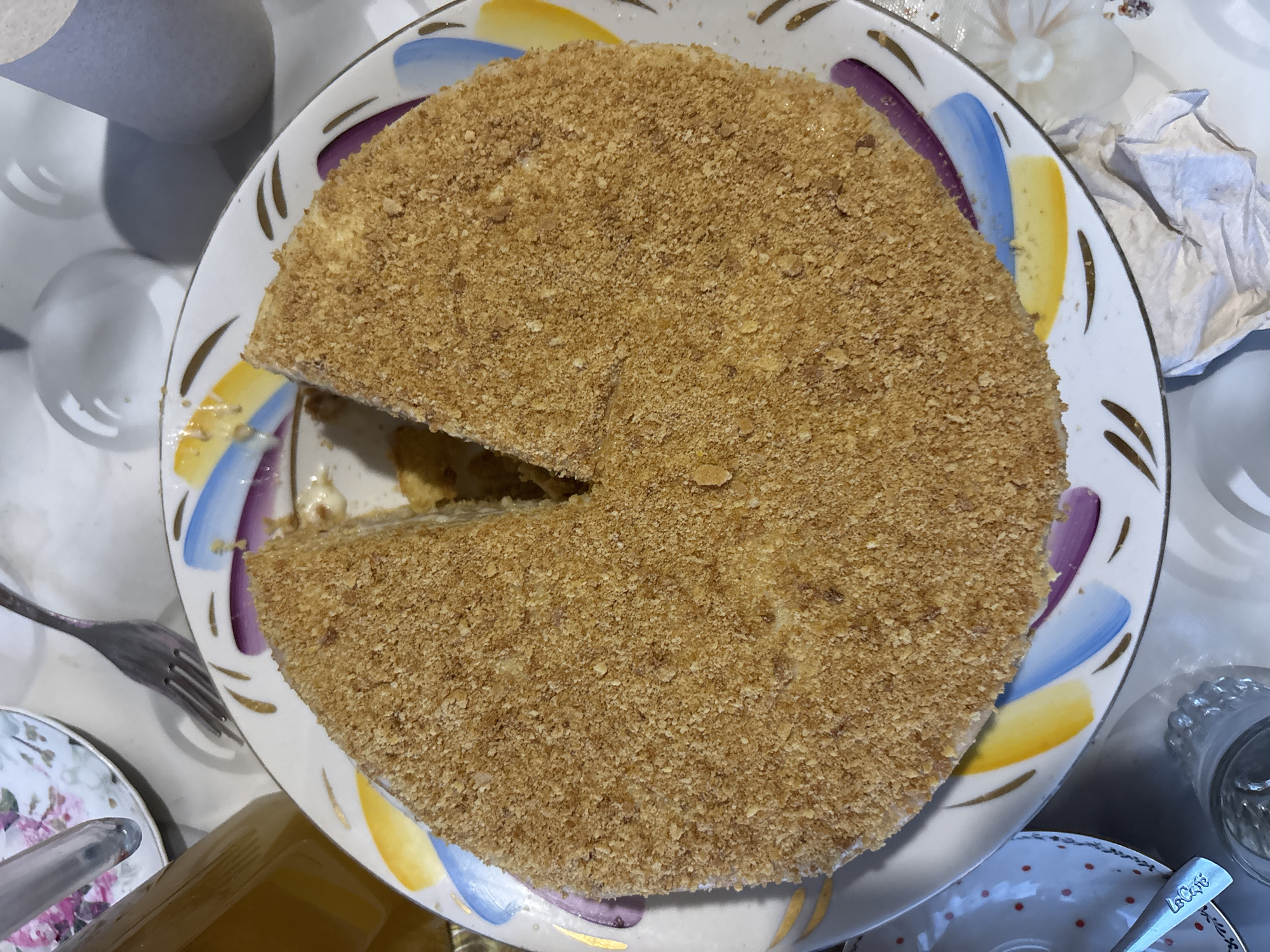 A top-down shot captures a partially consumed round cake on a decorative plate. The cake is light brown and covered in crumbs. A slice has been removed. The plate has a white background with a design featuring blue, yellow, and purple strokes. Surrounding the plate are various objects, including a fork, a cup with a floral pattern, a bottle, and a crumpled tissue. The setting is a table, possibly indoors. The location is Zagatuy, Russia.