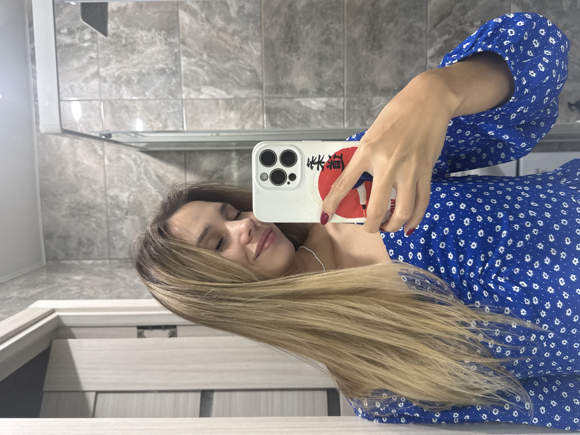 A young woman with long blonde hair takes a selfie in a bathroom. She is wearing a blue dress with a white floral pattern. Her eyes are closed and she is smiling. She is holding a white smartphone in her right hand. The bathroom has grey tiled walls and a glass shower enclosure. The image is a mirror reflection. The location is Chaikovsky, Russia.