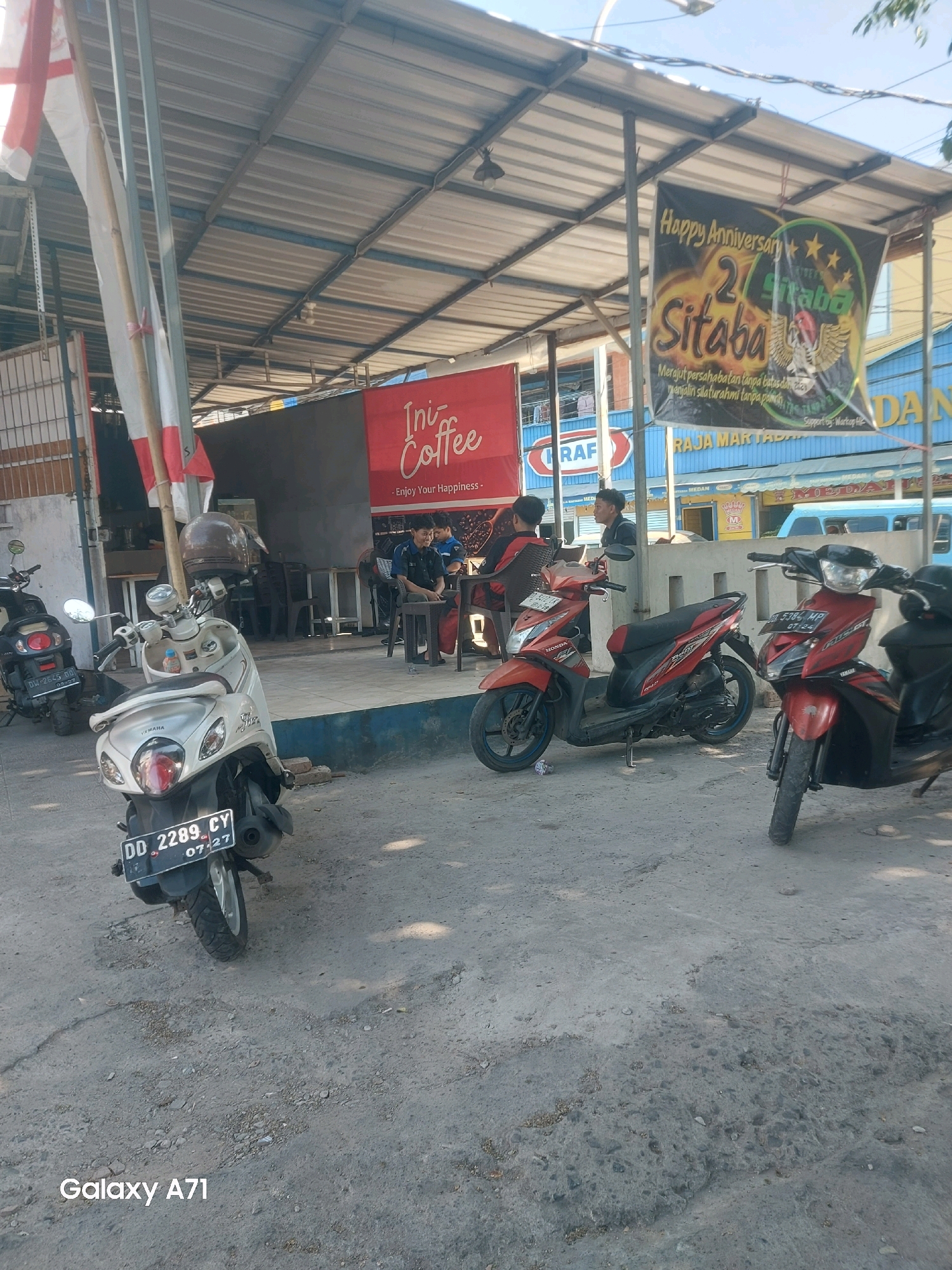 The image shows the exterior of a coffee shop in Makassar, Indonesia, likely on a sunny day. Several motorcycles are parked in front of the establishment. A banner hanging from the awning advertises a "Happy Anniversary 2 Sitaba" event, featuring graphics of wings and stars. Another red banner above the coffee shop entrance displays "Ini Coffee" and the tagline "Enjoy Your Happiness." Inside the shop, two young men in dark shirts are seated at a table, and another man in a blue shirt is also visible. The ground is paved with rough concrete, showing signs of wear and tear. The overall atmosphere suggests a casual, outdoor dining or gathering space.
