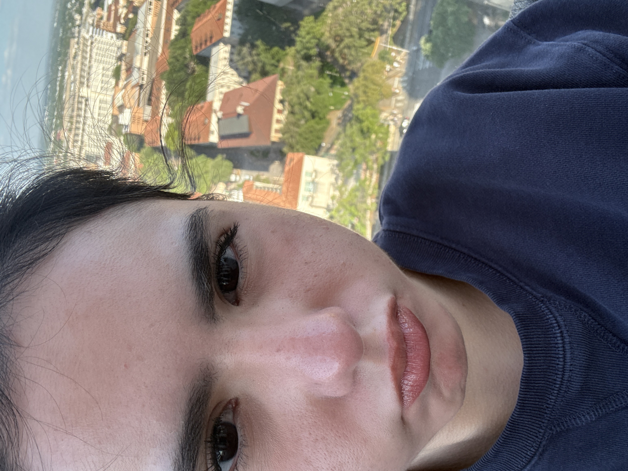A close-up selfie of a person with dark hair and brown eyes, wearing a dark blue sweatshirt. The background shows a city landscape with buildings and trees. Based on the architectural style and the layout of the buildings, it is likely taken from a high point in Austin, Texas, United States. The orientation of the image is rotated, showing the person at an angle.