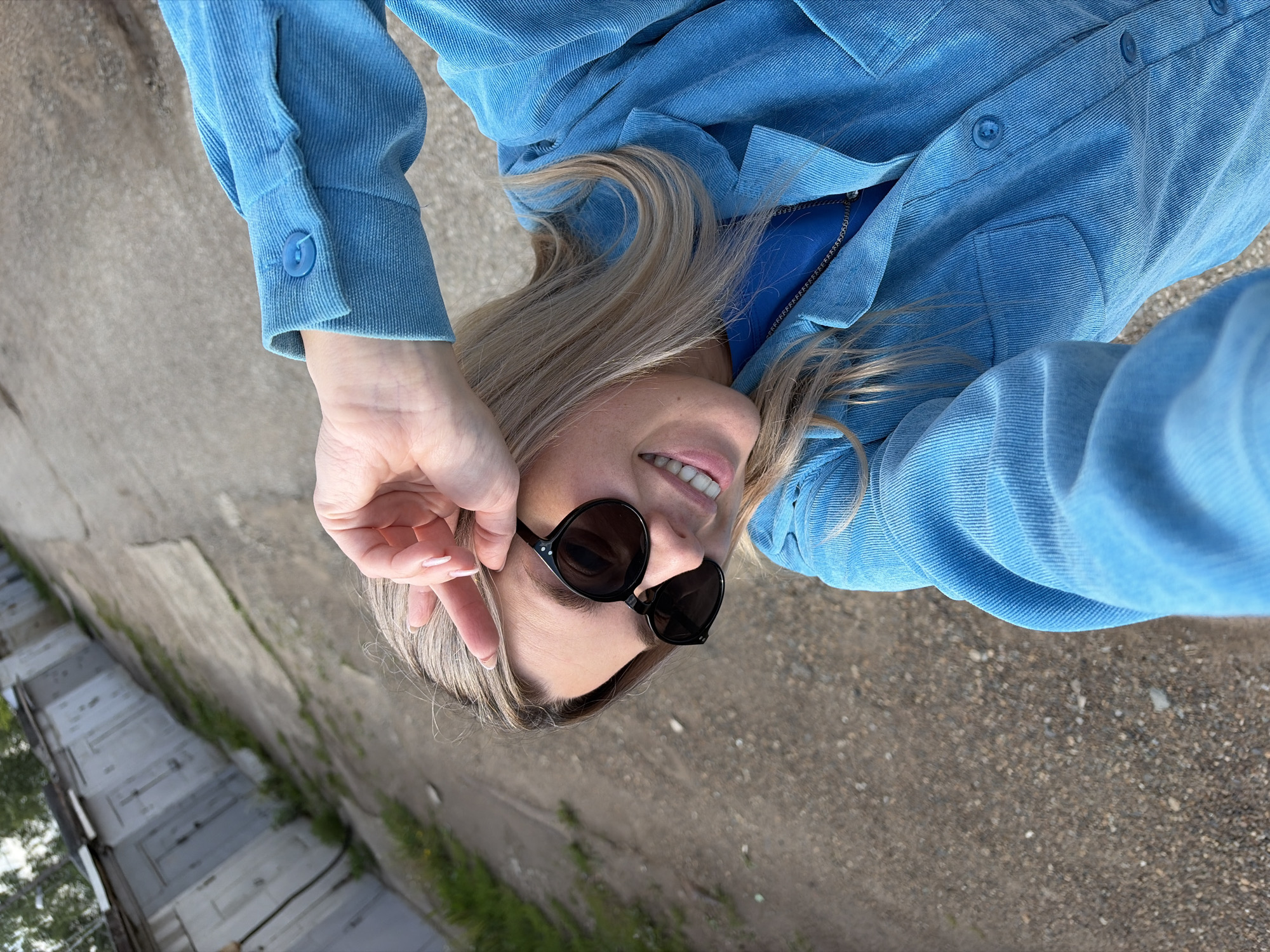 An overhead portrait captures a blonde-haired individual, likely female, lying outdoors on a paved surface. The image is taken from above, with the subject positioned diagonally across the frame. She wears round, dark-lensed sunglasses and a light blue corduroy shirt or jacket, which is partially unzipped to reveal a darker blue garment underneath. Her left hand, adorned with light-colored nail polish, is raised towards her face, appearing to adjust her sunglasses or touch her hair. A cheerful smile is visible, with her mouth slightly open.
The background primarily consists of grey asphalt or gravel. To the left side of the frame, there is a concrete wall or fence line with patches of green vegetation, and further back, several grey, rectangular structures resembling garages or storage units are visible. The bright, clear lighting suggests it is daytime. The scene evokes a relaxed, casual mood. The setting, particularly the garage-like structures and paved ground, is consistent with an urban or semi-industrial area, located in Chaikovsky, Russia. No visible text is present.