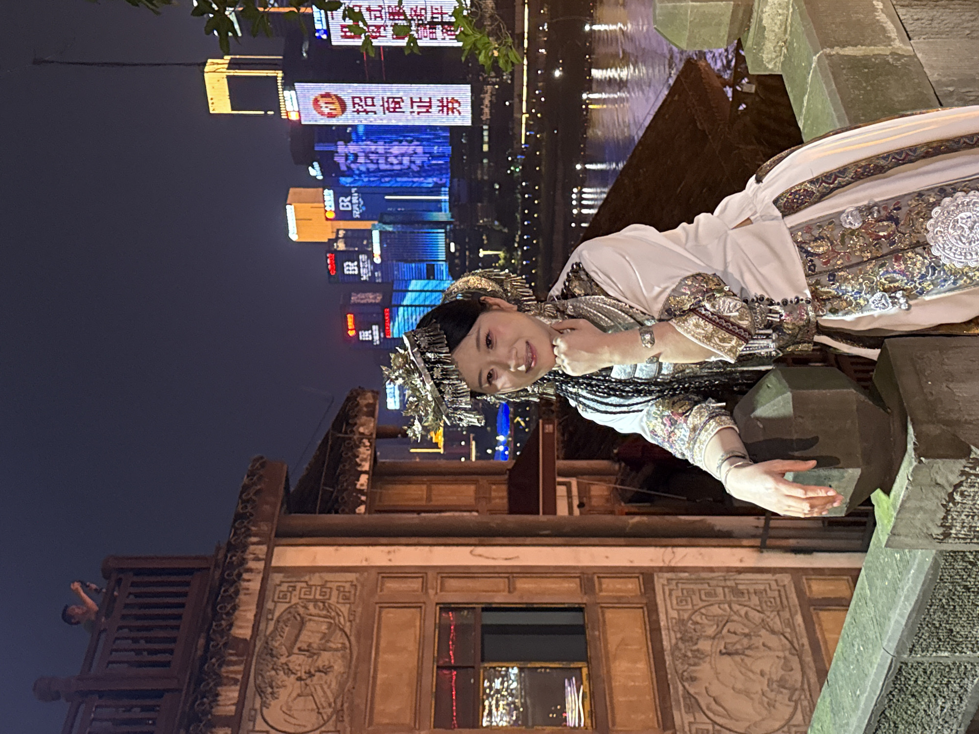 A woman wearing an ornate traditional costume with silver headwear and jewelry poses on a stone bridge at night. The background features a cityscape with illuminated skyscrapers. The bridge is constructed with traditional architectural elements, including decorated panels. A person is visible on a structure adjacent to the bridge. The location is identifiable as Yuzhong District, China, based on the distinctive architectural style and cityscape.