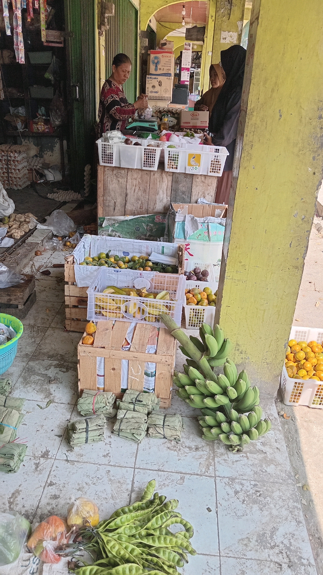 In Kunduran, Indonesia, a market stall is captured from a high-angle perspective. A woman in a red and black patterned top is weighing produce on a green digital scale. Beside her, two other women, one in a brown hijab and the other in a black hijab, are standing and looking towards the stall. The stall displays an array of fruits and vegetables, including oranges, bananas, starfruit, and what appear to be packaged food items wrapped in leaves. Crates and baskets are used to organize the produce. In the foreground, bundles of green beans and other vegetables are laid out on the tiled ground. The background shows a building with a yellow facade and a green shuttered entrance. The scene suggests a busy marketplace environment.