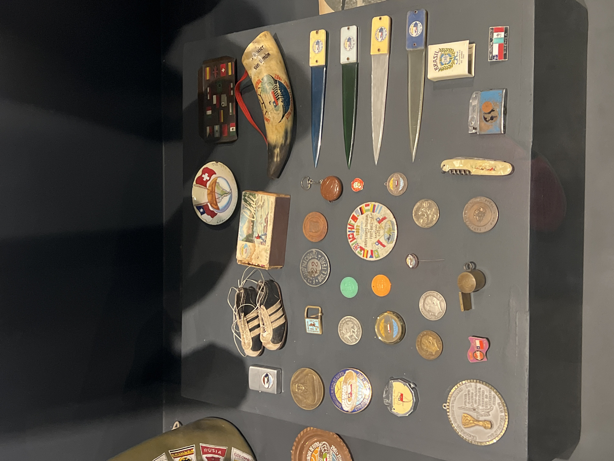 The image shows a collection of items mounted on a gray wall, likely a display or a memory board. The items appear to be souvenirs or memorabilia related to Chile and the 1962 FIFA World Cup, which was held in Chile.
Objects include:
- A decorative horn with lettering related to the 1962 World Cup.
- Four letter openers with matching handles.
- A wooden box and a matchbox with "Brasil" printed on it.
- Several coins and badges.
- A small pair of black and white soccer shoes.
- A pocket knife.
- A whistle.
- A medal with imagery of the FIFA trophy.
- A decorative plate with national flags.
- A small lighter.
- Flags.
The presence of the items relating to the 1962 FIFA World Cup, including a poster promoting the city of Arica, and Chilean themed souvenirs indicate this collection likely belongs to someone who has an association with that event, or with Chile.