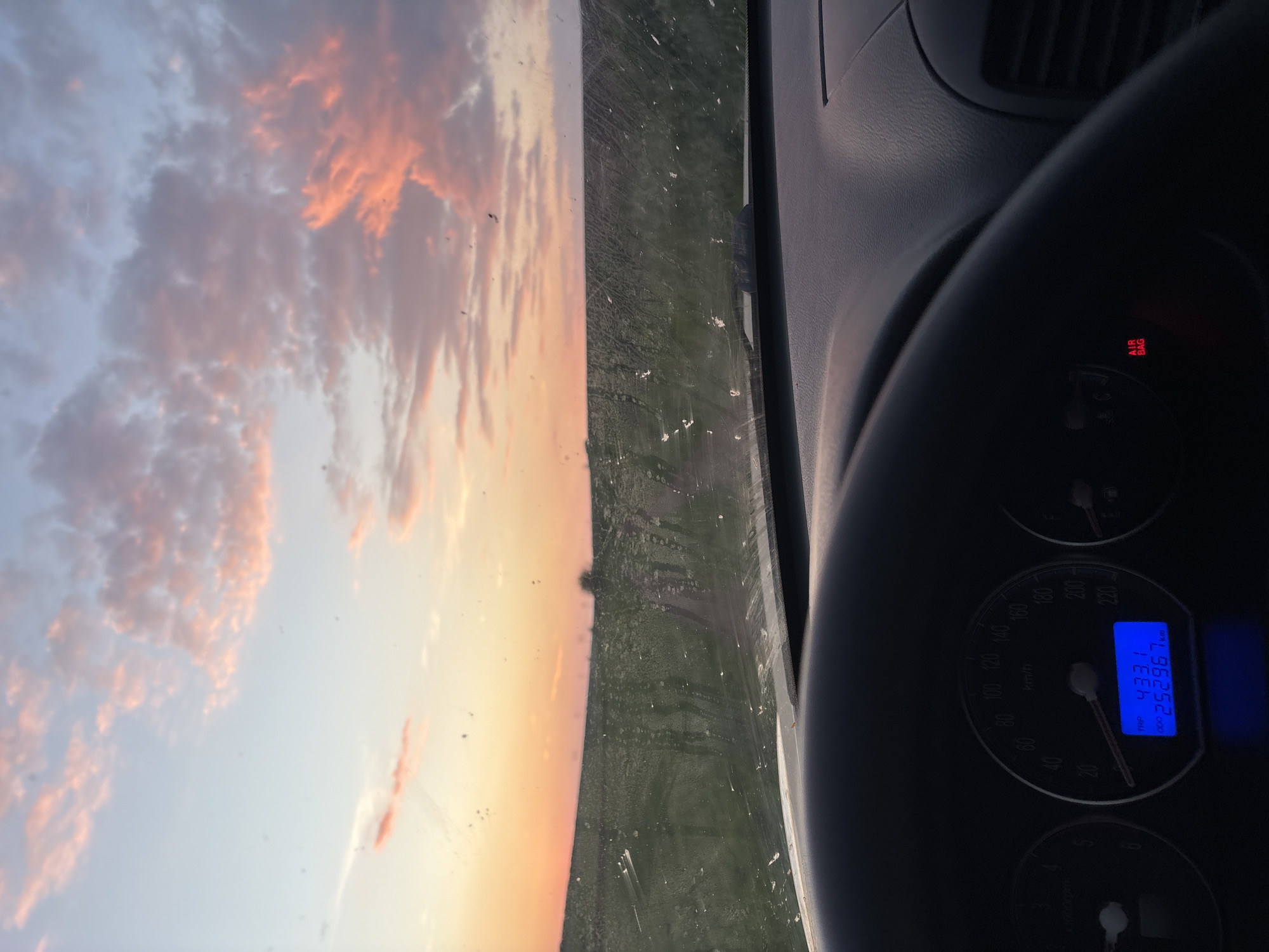 The image shows a view from inside a car, likely while driving. The dashboard and steering wheel are visible on the right side of the frame. The speedometer reads 433.1 for the trip and 252967km total. The "Air Bag" indicator light is illuminated. Through the windshield, a landscape is visible under a sky with pink-tinted clouds. The scene suggests a rural setting with an open field. The specific location is Lehchevo, Bulgaria.