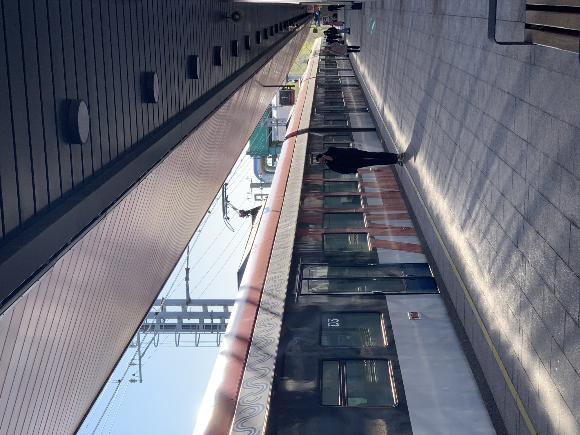 The image captures a train station platform in Moscow, Russia. A passenger stands alongside a train, facing away from the camera, appearing to look down at their phone. Other indistinct people are seen further down the platform. The platform is tiled and runs parallel to the train. A yellow safety line runs along the platform edge. An overhang structure is above the platform and the train. Overhead electrical lines are visible. The train's exterior has a color gradient from dark to light and a repeating blue design element. The number "03" is visible on the side of the train car.