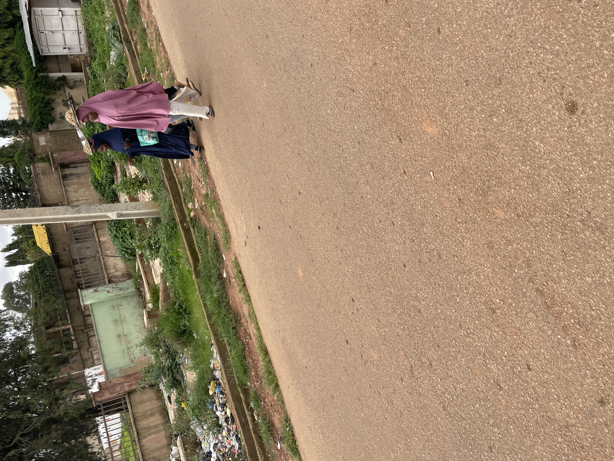 In Jos, Nigeria, two women walk on a paved road. The woman on the right wears a pink hijab and white pants, carrying a flat tray balanced on her head. The woman on the left wears a dark blue hijab and a bag with a patterned design slung over her shoulder. Behind them, a roadside ditch is filled with accumulated garbage and debris, bordered by a low concrete wall and overgrown grass. In the background, a fence separates the road from a property with a green wall, trees, and buildings. A white metal container is visible on the right side of the background. The sky is overcast.