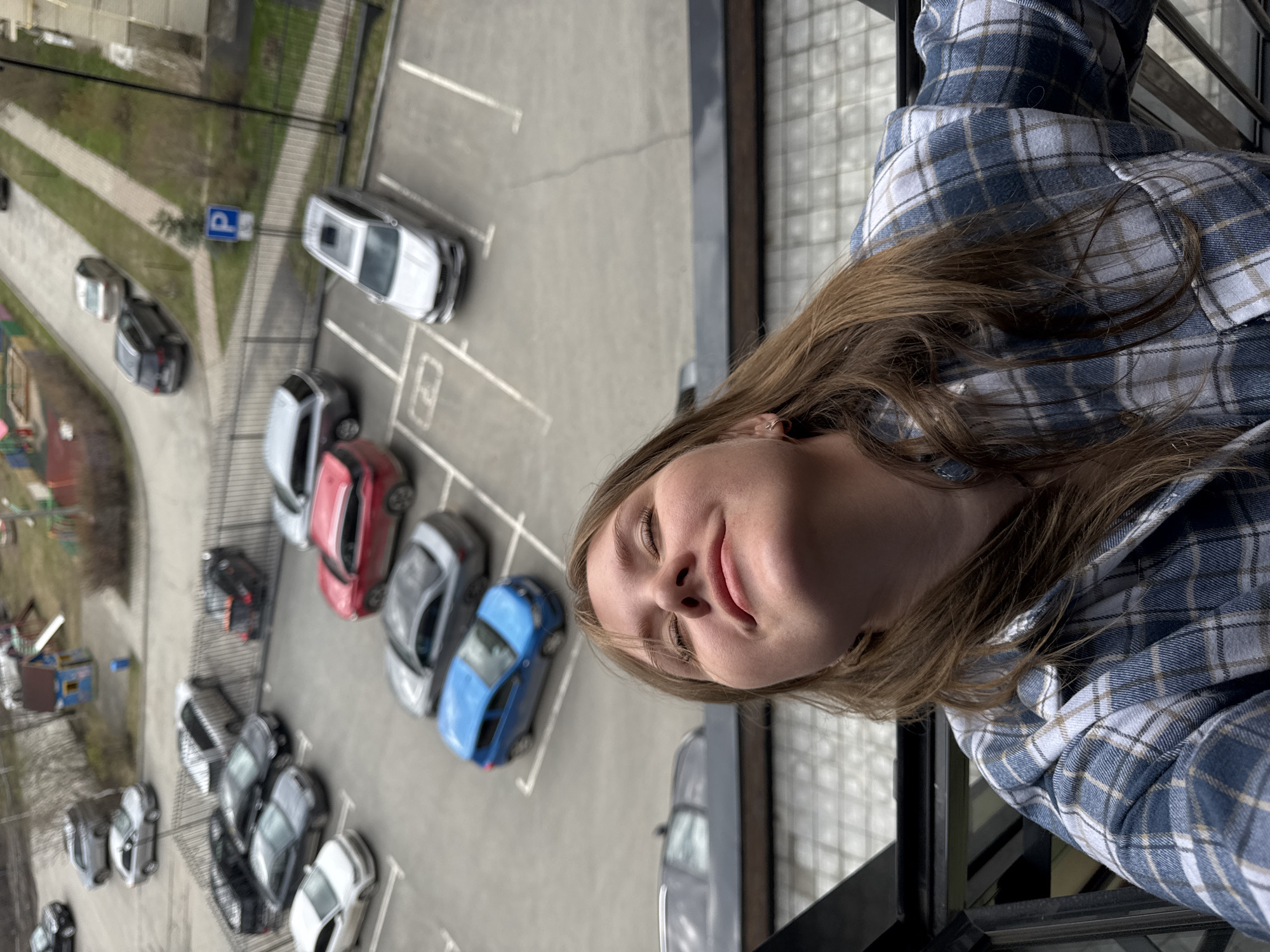 A young woman is featured prominently in the foreground of this image, captured from an elevated perspective, possibly from a balcony or high window. The image is rotated 90 degrees counter-clockwise from standard orientation.
The woman has long, light brown hair and is wearing a blue, white, and tan plaid flannel shirt. Her head is tilted slightly, and she has a relaxed expression with her eyes nearly closed and a gentle smile or smirk on her face. A small, simple hoop earring is visible in her left ear. A black metal railing, likely part of a balcony, frames the bottom right portion of the image.
The background, viewed from above, reveals a sizable asphalt parking lot with numerous cars of various colors (white, black, red, grey, blue) parked in clearly marked spaces. Adjacent to the parking area is a paved pathway bordered by green grass and some trees. A blue square sign with a white "P" (parking symbol) is visible near the pathway. In the distance, elements of a residential or urban environment, including what appears to be a children's play area, are discernible.
The scene is illuminated by natural daylight, indicating it is daytime, possibly under an overcast sky given the soft, even lighting. The woman's expression suggests a mood of contentment or relaxation. The context provided indicates the location is Perm, Russia.