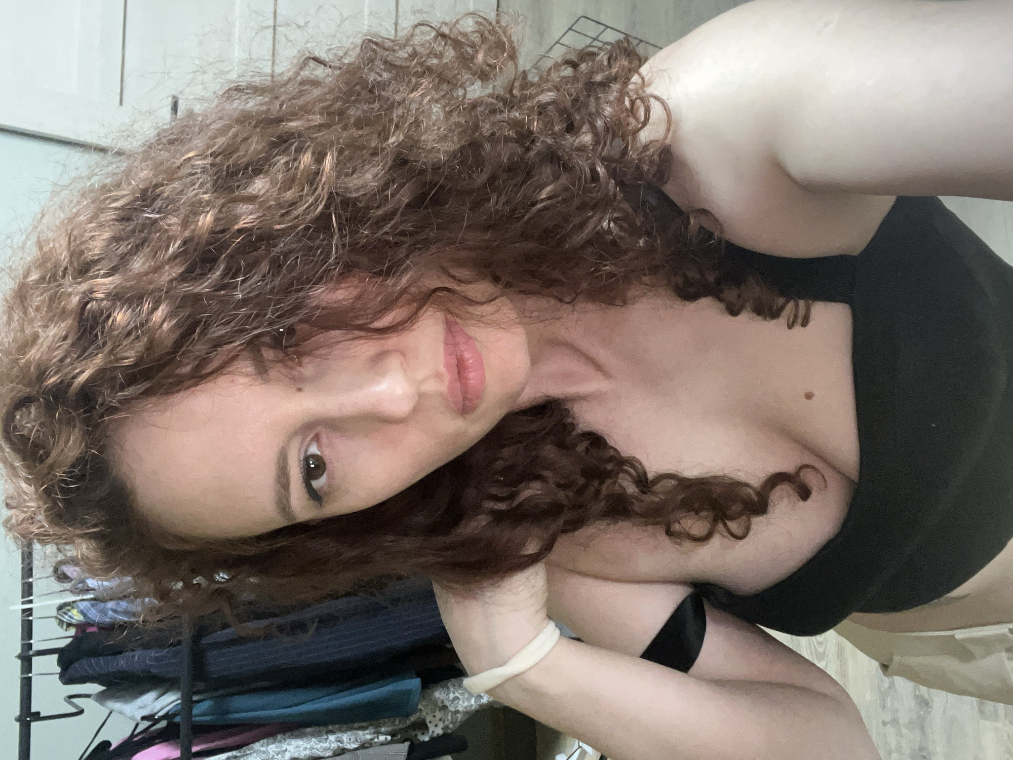 A close-up selfie shows a woman with voluminous, curly brown hair, partially framing her face. The image is oriented diagonally, with the woman looking directly at the camera with a neutral expression. Her brown eyes are subtly made up with eyeliner, and her lips have a soft pink tint. She has fair skin, and a small mole is visible on her upper chest.
She is wearing a black top or bra, and a light-colored, possibly white, wristband is on her left wrist. Her left arm is bent, with her hand near her hair on the left side of the frame. The background on the left reveals a black metal clothes rack or storage unit holding folded garments in dark and muted tones. The background on the right is a plain, light-colored wall. The lighting is soft and consistent with an indoor environment, offering no specific cues regarding the time of day or outdoor weather. The image itself contains no visible text or explicit geographical identifiers for Saint Petersburg, Russia, though that location is provided as context.