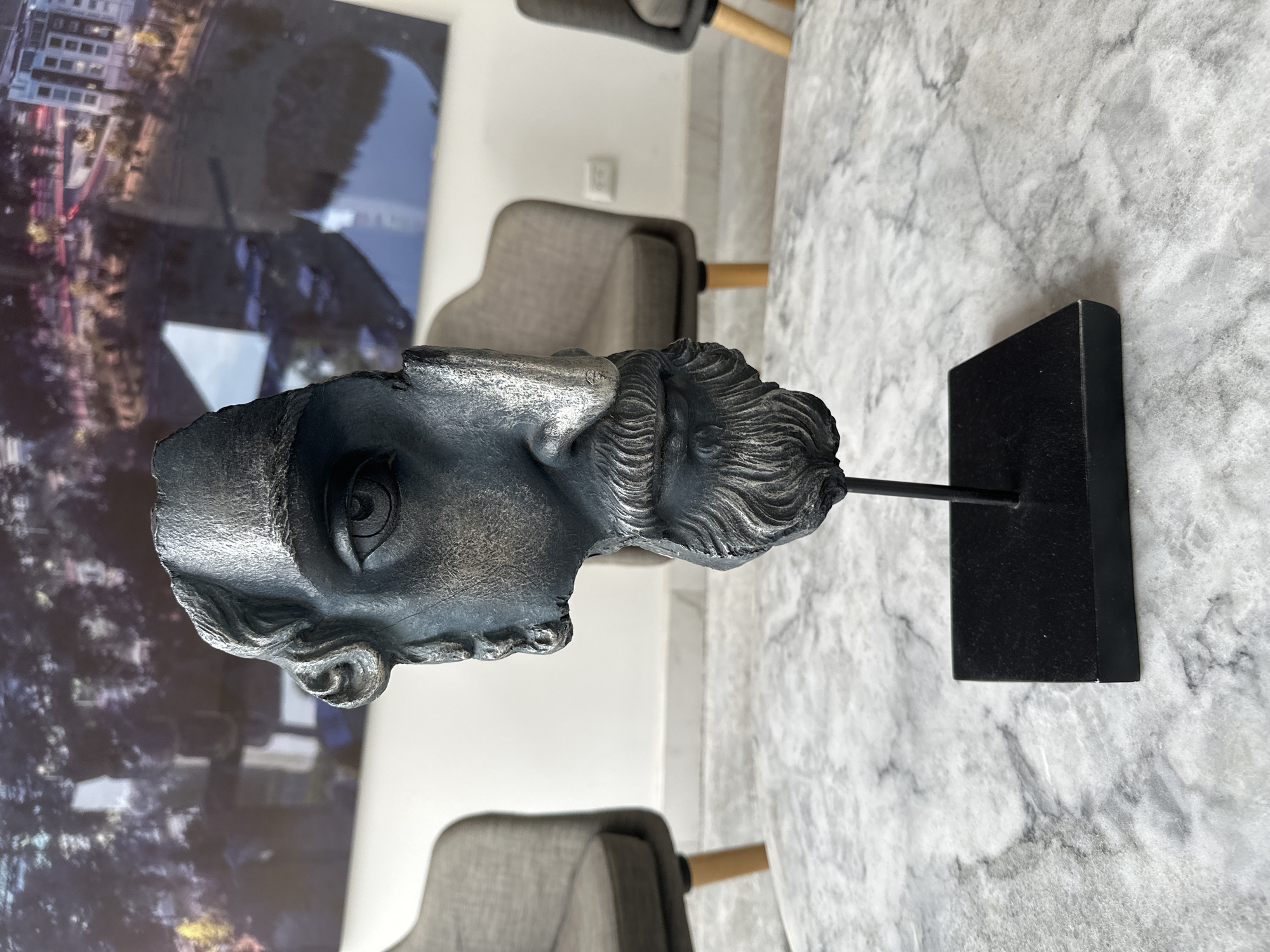The image shows a bust sculpture of a classical figure, seemingly Roman or Greek, resting on a black stand placed on a marbled surface. The sculpture is silver or gray in color, depicting a male head with detailed hair and facial features. The marbled surface is predominantly gray and white. In the background, out of focus, are furniture pieces like gray chairs with wooden legs, a wall, and a photograph or artwork depicting buildings and urban scenery. The image does not directly provide information about the location in the city of Puebla, Mexico.