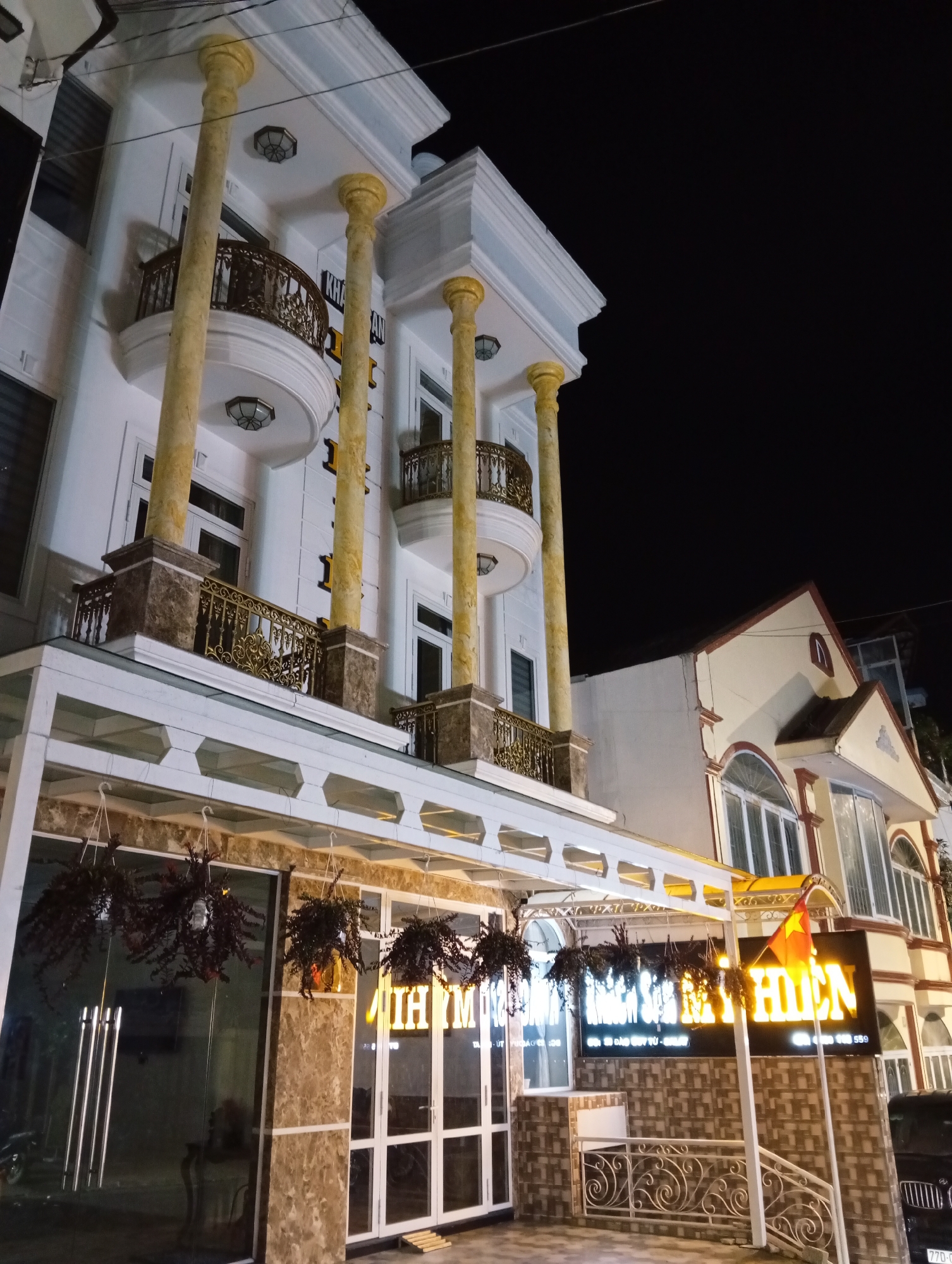 A photograph taken at night depicts the facade of a building in Da Lat, Vietnam. The building features a two-story structure with architectural details like columns, balconies with ornate railings, and a prominent sign illuminated with the words "NHA HANG SOM MAI HIEN." Hanging planters decorate the entrance. The image also shows a portion of a neighboring building with arched windows and a Vietnamese flag. A car is partially visible on the right side.