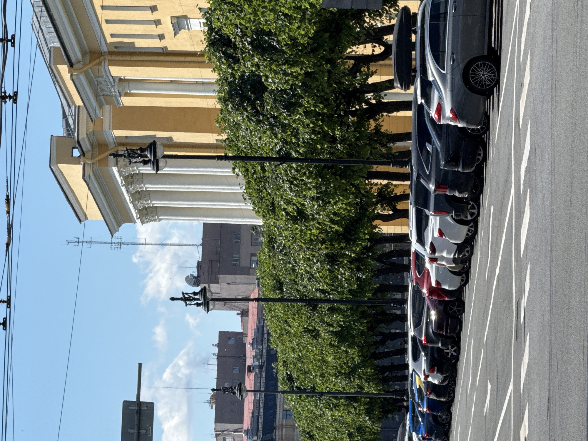 This image captures a street scene in Saint Petersburg, Russia, likely during daylight hours given the bright illumination and clear blue sky with scattered clouds. The foreground displays a row of parked cars of various makes and models, mostly SUVs and sedans, lining a street with visible lane markings. The cars are predominantly modern and appear to be in good condition.
Behind the parked cars, a line of lush green trees with dense foliage forms a natural barrier. Beyond the trees, an elegant, yellow building with classical architectural elements, including large white columns and ornate cornices, dominates the background. The building's facade suggests a significant institutional or historical structure. In the distance, other urban buildings and infrastructure, including a tall communication tower, are visible against the sky.
Two traditional lampposts with elaborate designs stand along the street, spaced between the trees, contributing to the urban aesthetic. The overall impression is a well-maintained city street with a blend of modern vehicles and historical architecture, set against a backdrop of greenery and a clear sky. There are no people visible in the immediate scene.