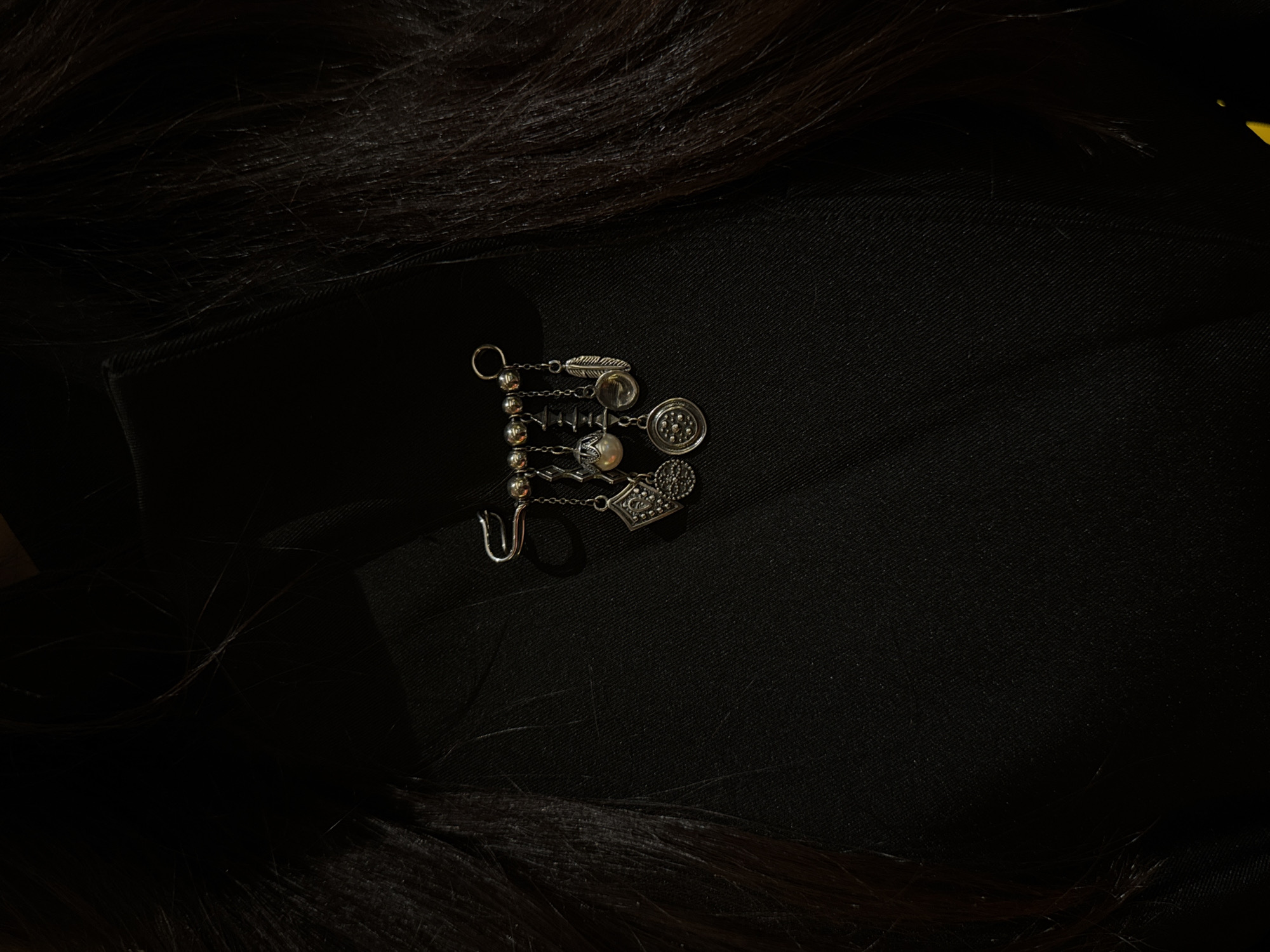 The image displays a silver-toned piece of jewelry, possibly an ear cuff, earring, or decorative brooch, centrally positioned on a dark black fabric surface. The jewelry consists of multiple small charms or pendants attached to a chain-like element adorned with small metallic beads. Visible charms include a feather shape, a spherical bead, a square element with an embossed design, a circular piece with a radiating pattern (possibly floral or geometric), and another distinct square-shaped charm. A U-shaped hook or clasp is present at one end of the jewelry.
Dark, natural hair frames the composition, appearing along the left side and bottom of the image, with individual strands visible, adding texture to the dark background. The black fabric beneath the jewelry appears to be a textile, possibly an item of clothing, exhibiting a subtle weave pattern. The lighting is subdued, highlighting the metallic sheen of the jewelry against the dark backdrop. The overall scene is a static, close-up shot, likely intended to showcase the jewelry. There is no discernible activity taking place, and no text is visible within the image. The setting is in Yerevan, Armenia.