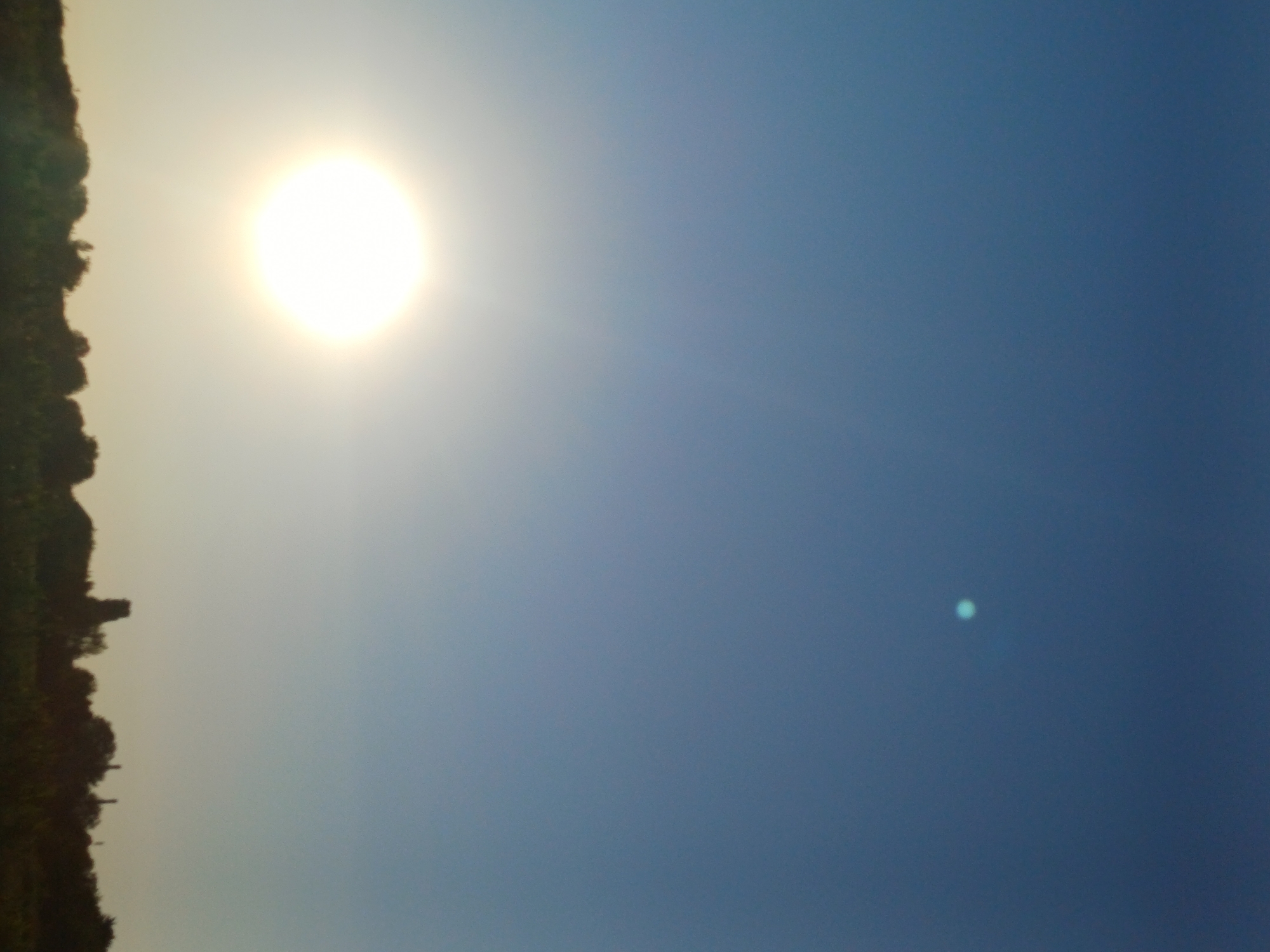 A vertical, low-resolution, outdoor photograph captures a bright sun partially visible on the left, with a faint, greenish-blue circular object in the upper right quadrant of the sky. The sky transitions from a bright blue at the top to a lighter, hazy tone near the horizon. A silhouetted treeline with some indistinct structures, possibly chimneys or buildings, forms the bottom edge of the image, suggesting a rural or suburban landscape. The specific location within Lutsk, Ukraine, cannot be determined from the visual information provided.