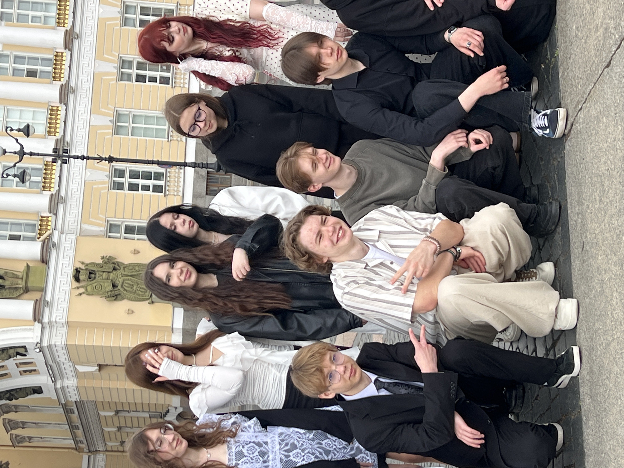A group of 11 young adults are arranged in a vertical, layered composition in front of the Winter Palace in Saint Petersburg, Russia. They are sitting close together, some with their arms around each other. Their clothing ranges from formal suits and dresses to casual hoodies and jeans. Some are looking directly at the camera. The background is the ornate yellow facade of the Winter Palace, with its detailed architectural elements like windows, columns, and statues.