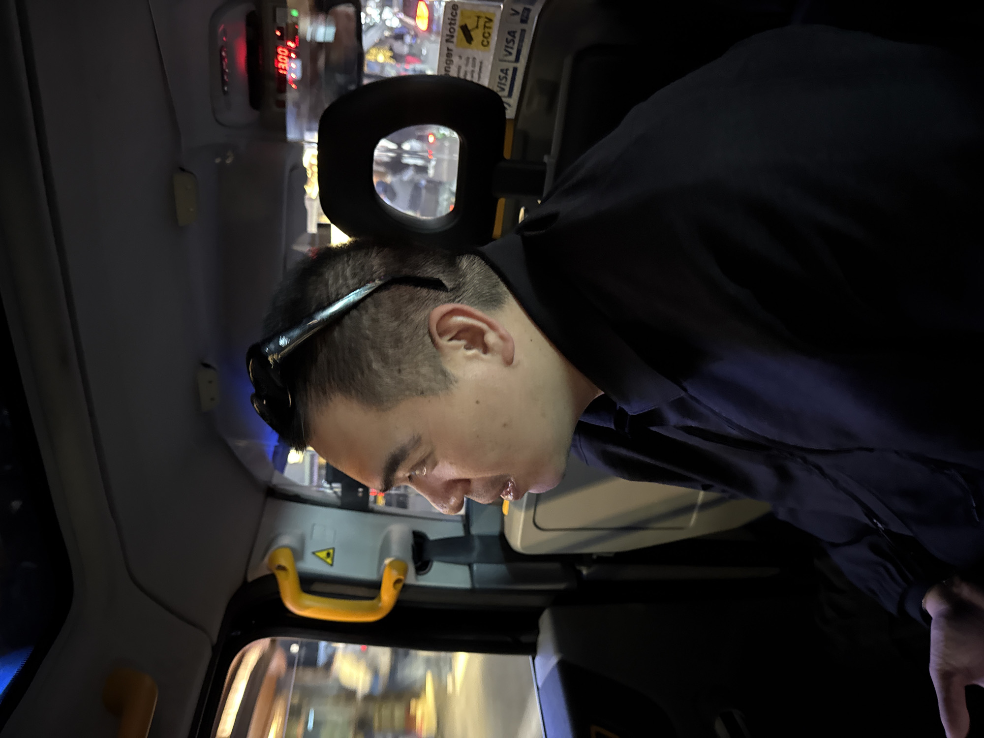 An Asian man with black sunglasses resting on his head is seated in a London black cab. The cab meter displays a fare of £13.00. Signage indicates acceptance of VISA payments and displays a CCTV notice. A yellow grab handle is visible on the partition. The interior is typical of London taxis, with a headrest in view. The background through the cab window shows blurred city lights, likely indicating a nighttime setting in London.