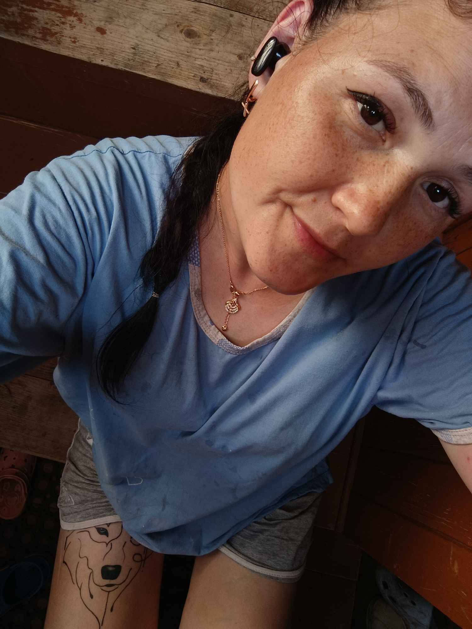 A close-up, high-angle shot shows a woman from the shoulders up, posing for a selfie. She is wearing a light blue t-shirt and grey shorts, with a tattoo of a wolf's head visible on her left thigh. A gold necklace with a key pendant rests on her chest. She has dark hair styled in a braid, and is wearing an earbud in her right ear. The background appears to be a wooden surface, possibly a porch or deck. The lighting suggests it is daytime. The exact location is unknown.