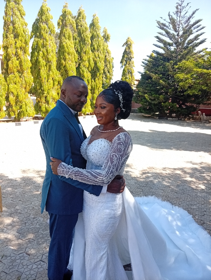 The image displays an adult male and female, consistent with a bride and groom, embracing outdoors under a clear blue sky. The male, on the left, wears a dark blue suit with a bow tie, his left arm around the female's waist as he looks down at her. The female, on the right, wears a white, elaborately beaded mermaid-style wedding gown featuring a sweetheart neckline, long sheer embellished sleeves, and a substantial train. Her hair is styled in a high updo with a decorative hairpiece, and she wears pearl jewelry. Her right arm is around the male's waist, and she is smiling broadly, looking slightly forward and to her left. They stand on a geometrically patterned paved surface. The background features a row of tall, slender evergreen trees with bright green foliage, and a larger, darker green evergreen tree to the right. A partial view of a building with reddish tones is visible in the distant background. The specific location within the city of Lankan, Nigeria, cannot be determined from the visual information present in the image.