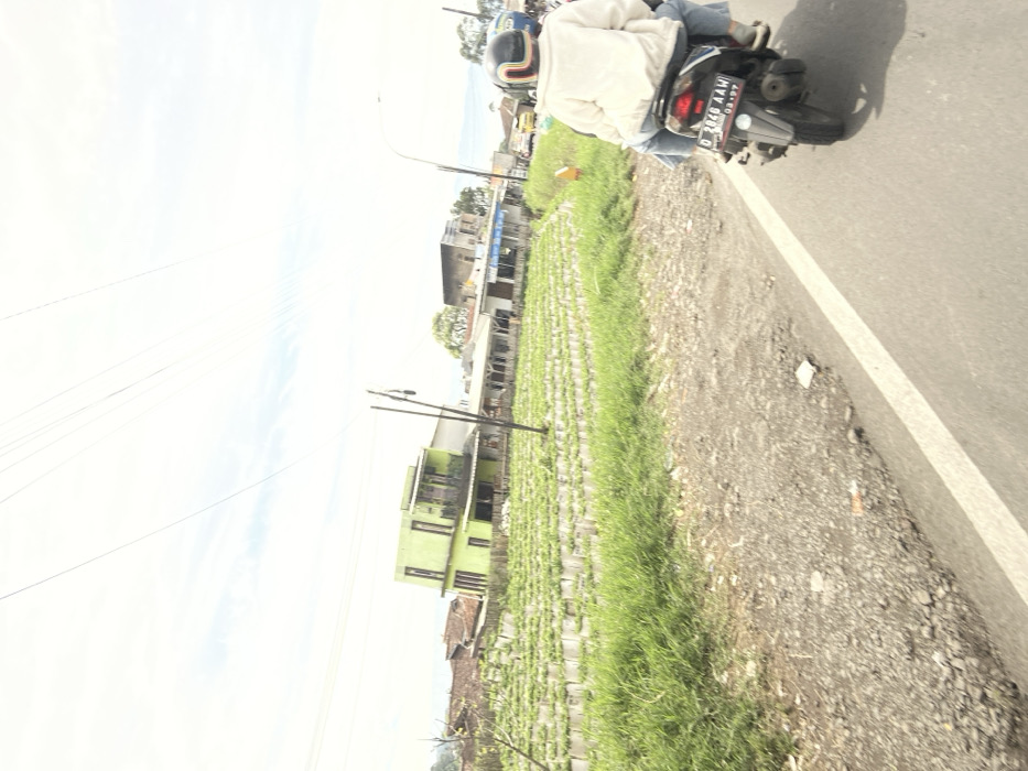 The scene is a road in Rancabali, Indonesia (latitude: -7.122, longitude: 107.428). A person is riding a motorcycle on the paved road, heading towards the camera. The road is bordered by a gravel shoulder and then by a field of plants in rows, possibly strawberry plants. Several buildings can be seen in the distance, including a light green two-story building on the left side of the frame. The sky is cloudy with a few visible power lines. There are trees in the background on both sides of the road, and some other vehicles can be seen traveling in the distance. The license plate on the motorcycle reads "D 2846 AAM". The road is relatively empty with no other vehicles in the immediate vicinity of the motorcycle. The weather appears to be sunny with clear skies.