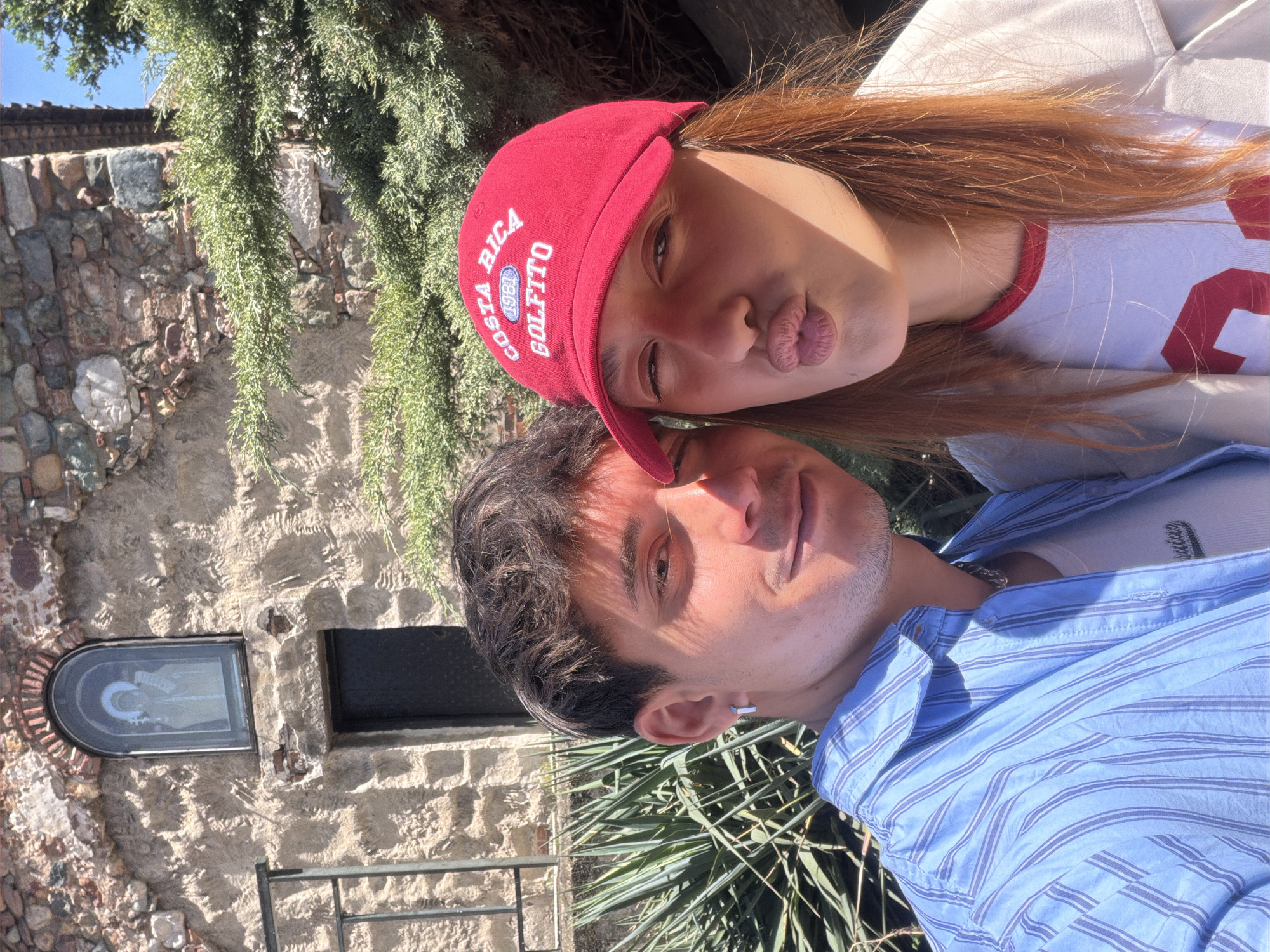 A photograph depicts two individuals in an outdoor setting. The foreground features a man wearing a light blue striped shirt, his head tilted slightly to the side. Above him is a woman with red hair wearing a white t-shirt with a red trim and a red cap with the words "Costa Rica Golfito 1981" embroidered on it. She is pursing her lips in a kissing gesture.
The background includes a stone wall with a small arched window displaying an icon. There is greenery present, suggesting a park or garden-like environment. The image appears to be taken in the city of Sofia, Bulgaria.