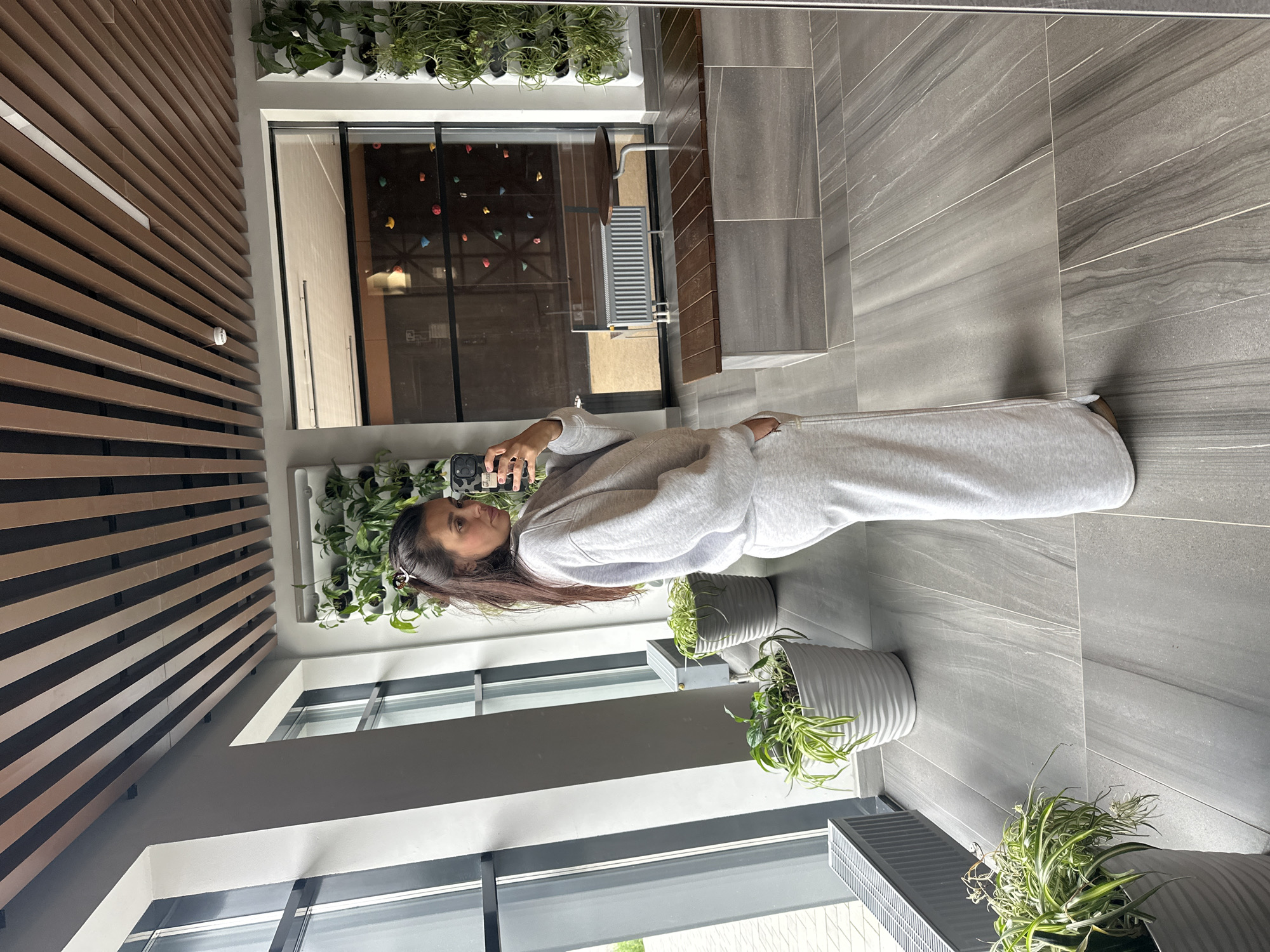 A full-body, eye-level, medium shot captures a young woman standing in a modern indoor space, potentially a lobby or communal area, while taking a mirror selfie. She is positioned in the foreground, facing left with her body slightly angled toward the camera, her head turned to look at her phone. She is wearing a light gray matching sweat suit, consisting of a long-sleeved sweatshirt and wide-leg pants. Her dark hair is pulled back loosely.
The environment features a polished concrete or large-format tile floor with a wood-grain pattern. To her left, large windows reveal an outdoor scene with some pavement and greenery, and a dark gray radiator is visible beneath the windows. Several potted plants with lush green foliage are strategically placed around the room, some in white ribbed planters. A vertical garden installation with many small plants is mounted on the wall behind her.
In the background, reflected in a large mirror that dominates the right side of the frame, we see more of the interior space. A wooden bench with metal legs is present, along with a small round table. Beyond this, a window looks into another room where colorful climbing holds are visible on a wall, suggesting a climbing gym or recreational facility. The ceiling is adorned with parallel wooden slats, with a continuous white light fixture running through them. The overall impression is one of contemporary design and a well-maintained, possibly urban, setting. There is no indication of the location being specifically in the city of Perm, Russia.